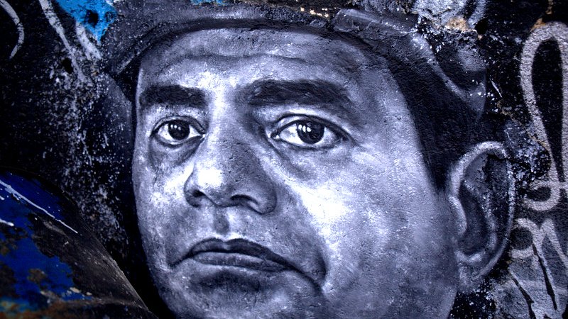 تظهر الصورة رسما جداريًا لوجه رجل، حيث يتميز التعبير بالعمق والتفاصيل الدقيقة. يُظهر الرسم عيونًا تعكس مشاعر قوية، والخطوط تتباين بين الظل والنور، مما يعطي إحساسًا بالحياة والواقعية. الخلفية تحتوي على ألوان داكنة ومخططات فنية أخرى، مما يبرز التركيز على وجه الرجل ويعزز من جمال العمل الفني.