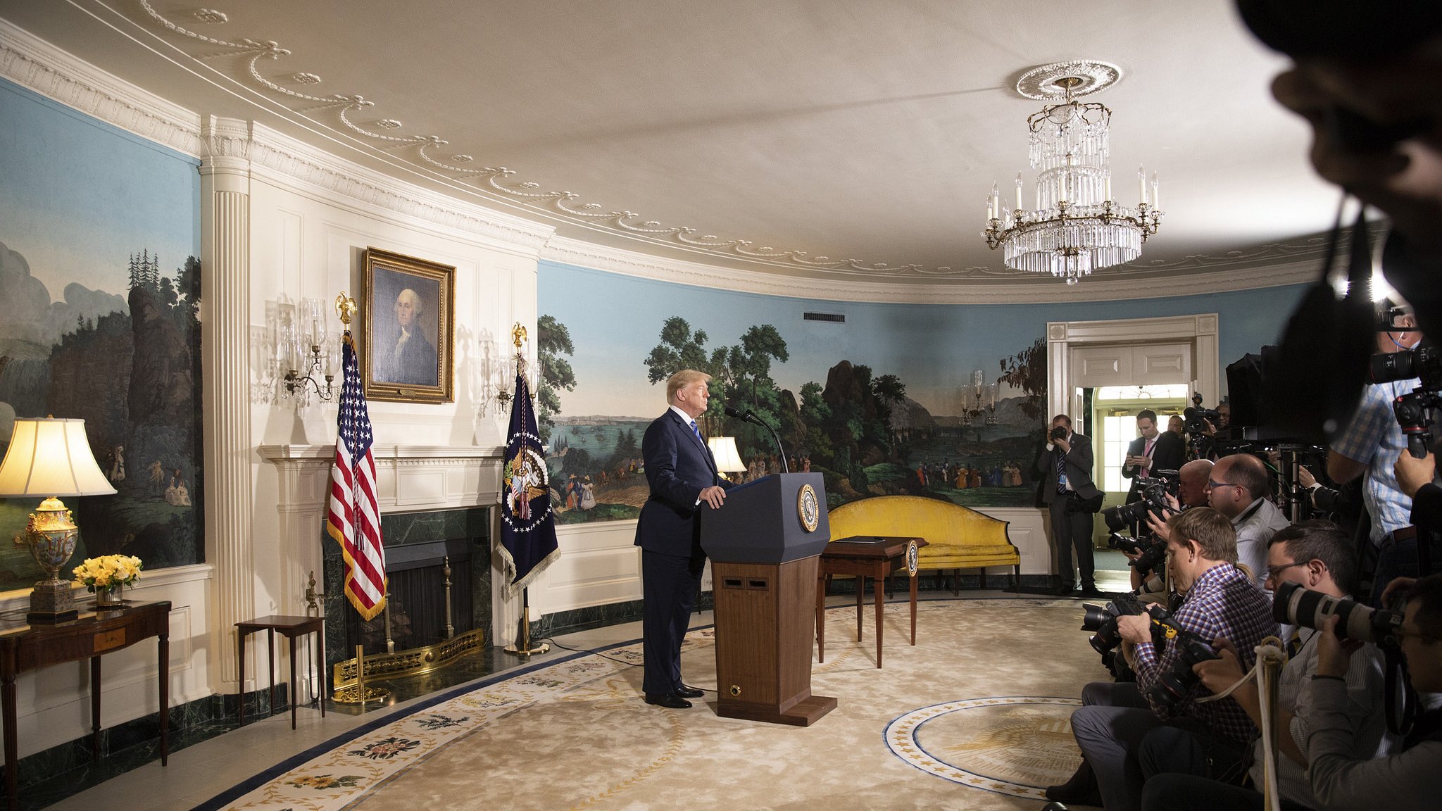 در این تصویر، دونالد ترامپ در حال سخنرانی است و در پس‌زمینه، دیواری با نقاشی‌های منظره و یک پرچم ایالات متحده و یک پرچم دیگر دیده می‌شود. او در کنار یک تریبون ایستاده و دوربین‌های خبرنگاران و عکاسان در جلو او قرار دارند. دکوراسیون اتاق زیبا و مجلل است، با یک فرش بزرگ و مجسمه‌هایی در اطراف. نور طبیعی از پنجره‌ها به داخل اتاق می‌تابد.