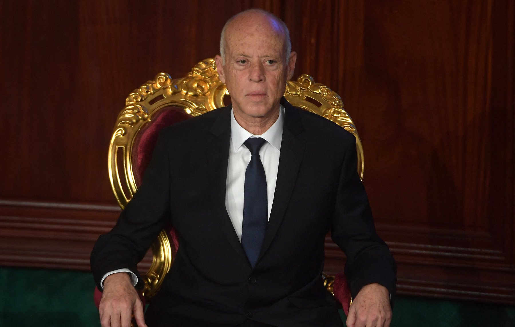 L'image montre un homme assis sur une grande chaise ornée, avec un décor en bois sombre en arrière-plan. Il porte un costume noir et une cravate sombre, affichant une expression sérieuse. Son attitude et son cadre suggèrent une ambiance officielle ou formelle.