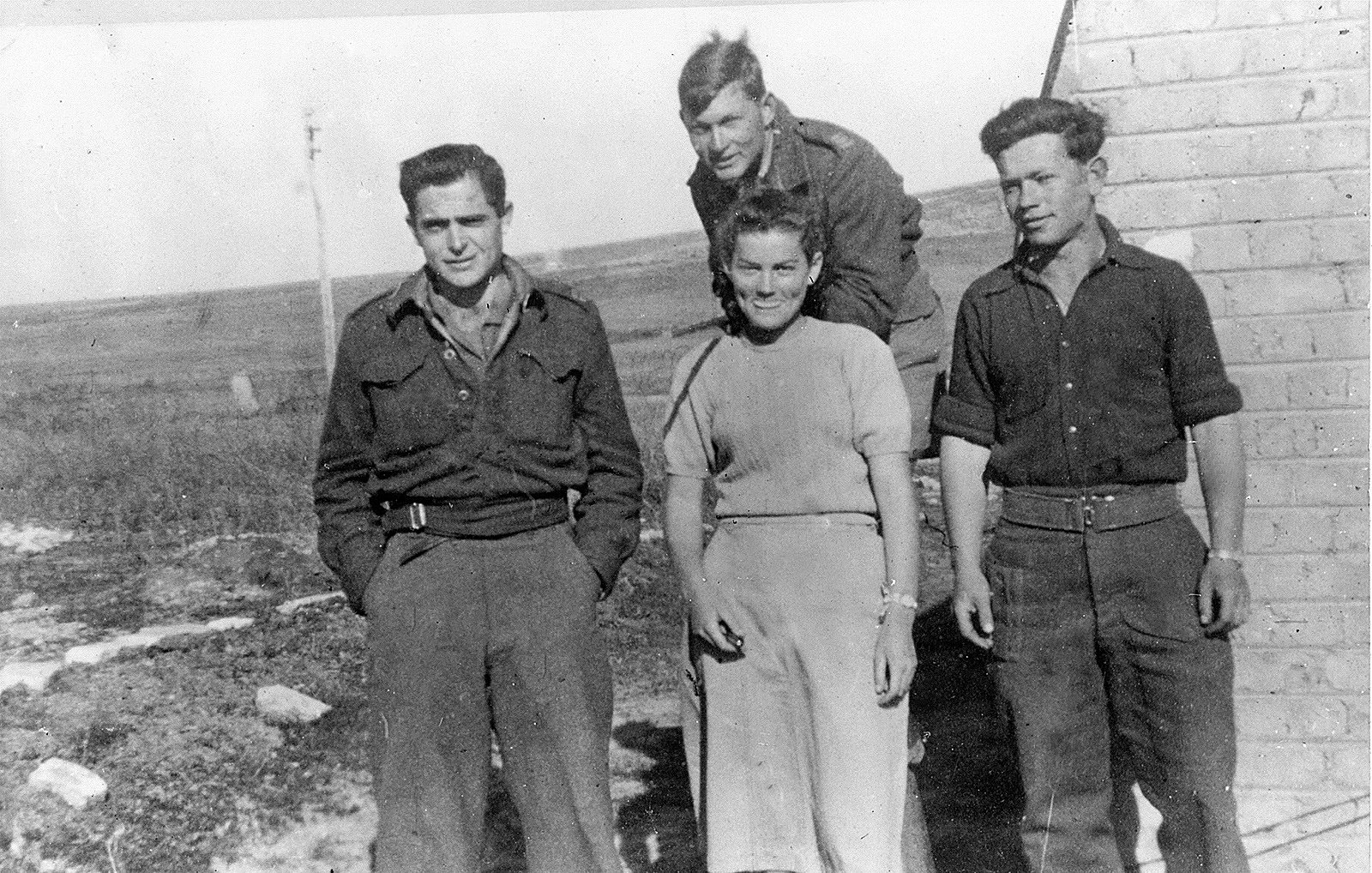 L'image montre quatre personnes se tenant debout à l'extérieur, probablement sur un terrain dégagé. Il y a trois hommes et une femme. Les hommes portent des uniformes militaires ou des vêtements utilitaires, tandis que la femme est habillée simplement dans une robe. L'environnement semble rural, avec une petite bâtisse en arrière-plan et des champs en toile de fond. Tout le monde affiche des expressions neutres ou souriantes, créant une atmosphère de camaraderie.