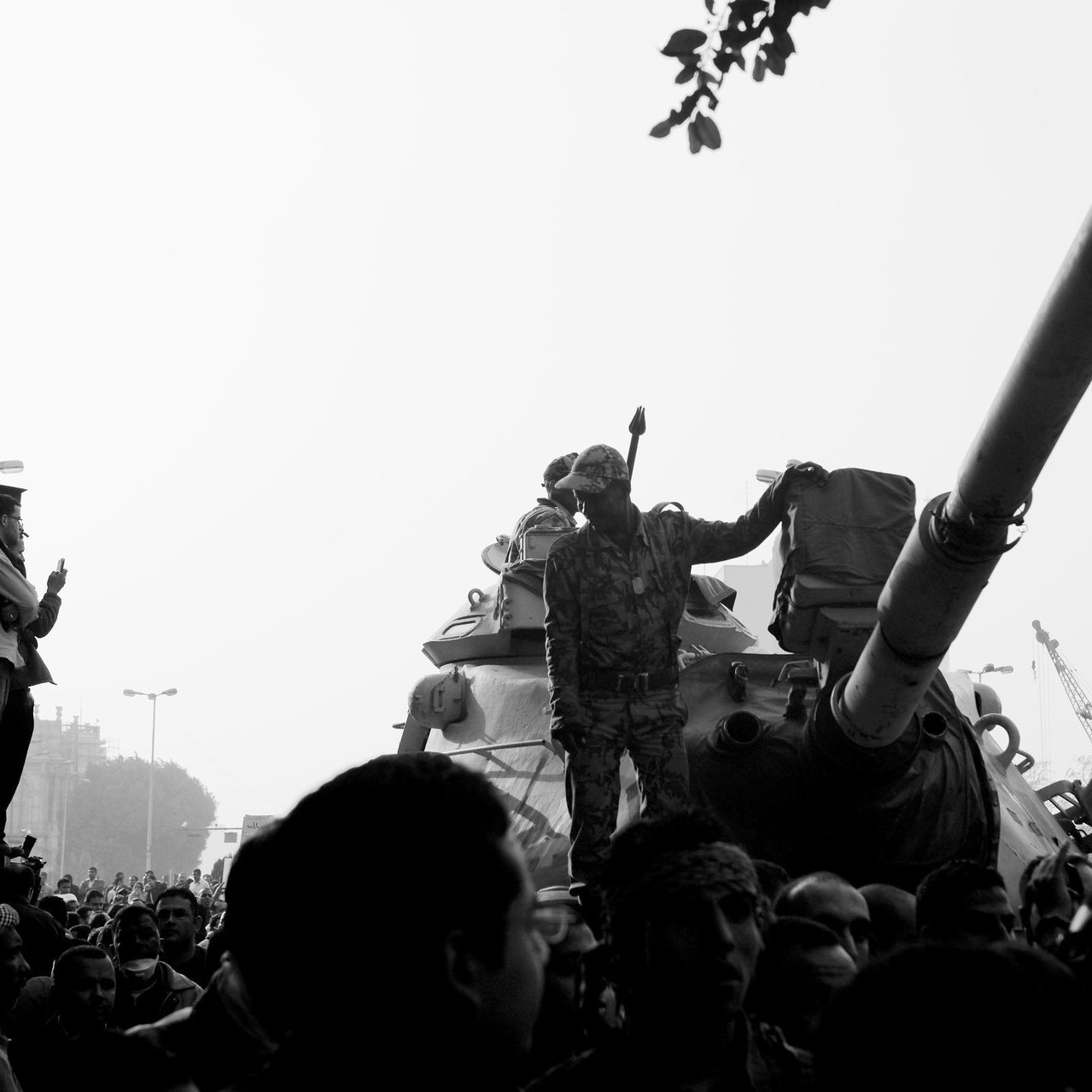 L'image montre une scène de manifestation ou de rassemblement avec une atmosphère intense. Au centre, on peut voir un soldat debout sur un char, levant le bras en signe de défi ou de puissance. Le char est entouré d'une foule dense de personnes, certaines levant les bras ou prenant des photos. L'arrière-plan semble urbain, avec des bâtiments et un lampadaire. La photo est en noir et blanc, ce qui ajoute à la dramaturgie et à la tension de la scène.