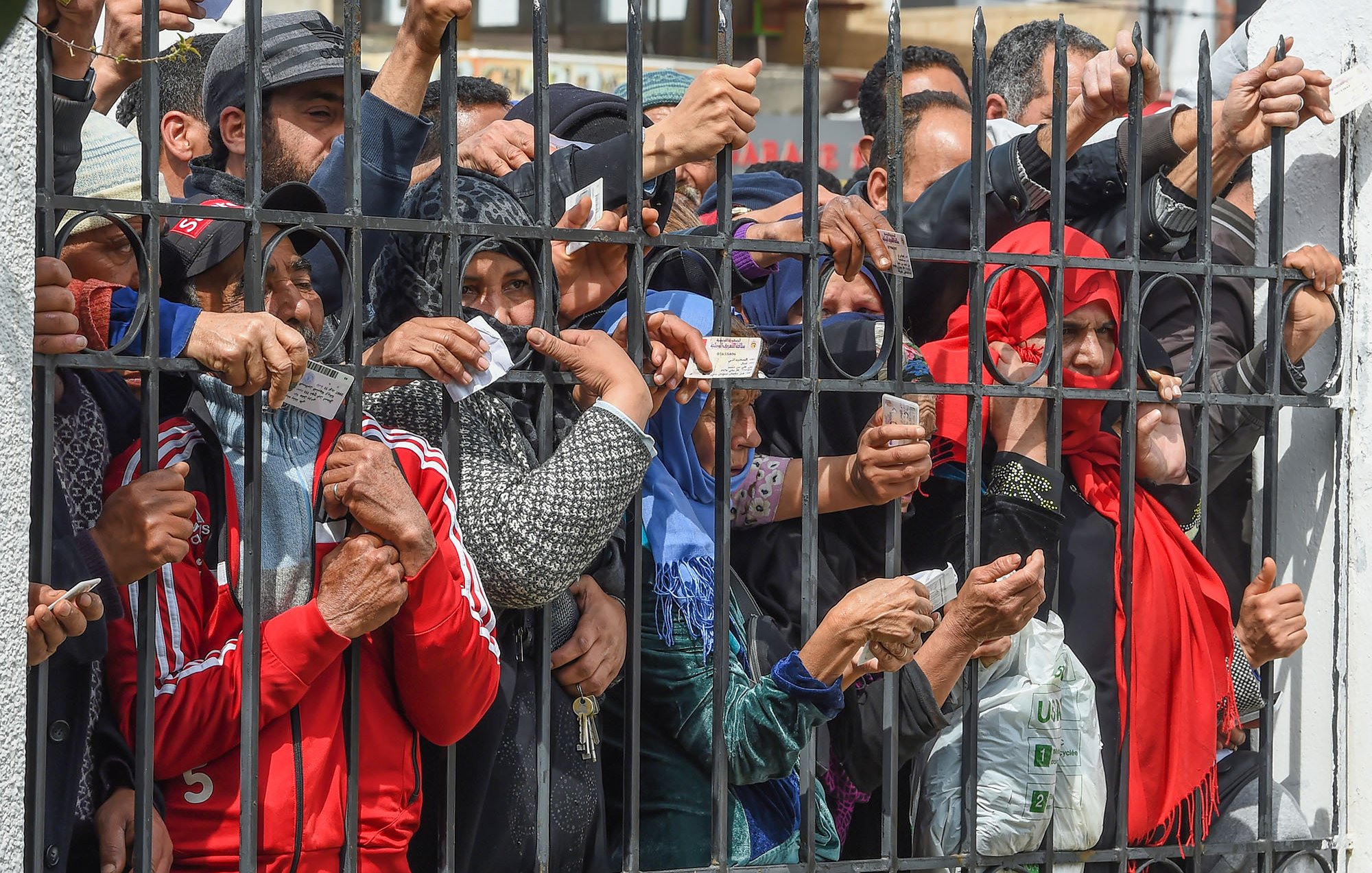 L'image montre un groupe de personnes rassemblées derrière des barreaux, semblant impatientes ou en attente. On peut voir des mains tendues à travers les barreaux, certains tenant des objets comme des cartes ou des papiers. Les visages de quelques personnes sont également visibles, exprimant diverses émotions. L'ambiance semble tendue, suggérant une situation de demande ou d'attente d'assistance. Les vêtements des individus montrent une diversité de styles, et il y a une impression de solennité dans la scène.