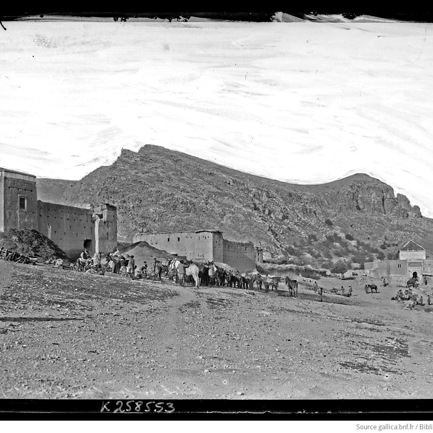 L'image présente un paysage désertique avec des montagnes en arrière-plan. À gauche, on aperçoit des bâtiments en pierre, probablement des fortifications ou des maisons, qui semblent très anciens. Au premier plan, un groupe de personnes est visible, accompagné de plusieurs animaux de charge, comme des chameaux ou des ânes. Le ciel est clair, montrant une atmosphère paisible, et le terrain est aride, typique des régions montagneuses désertiques. L'ensemble dégage une impression d'histoire et de culture.