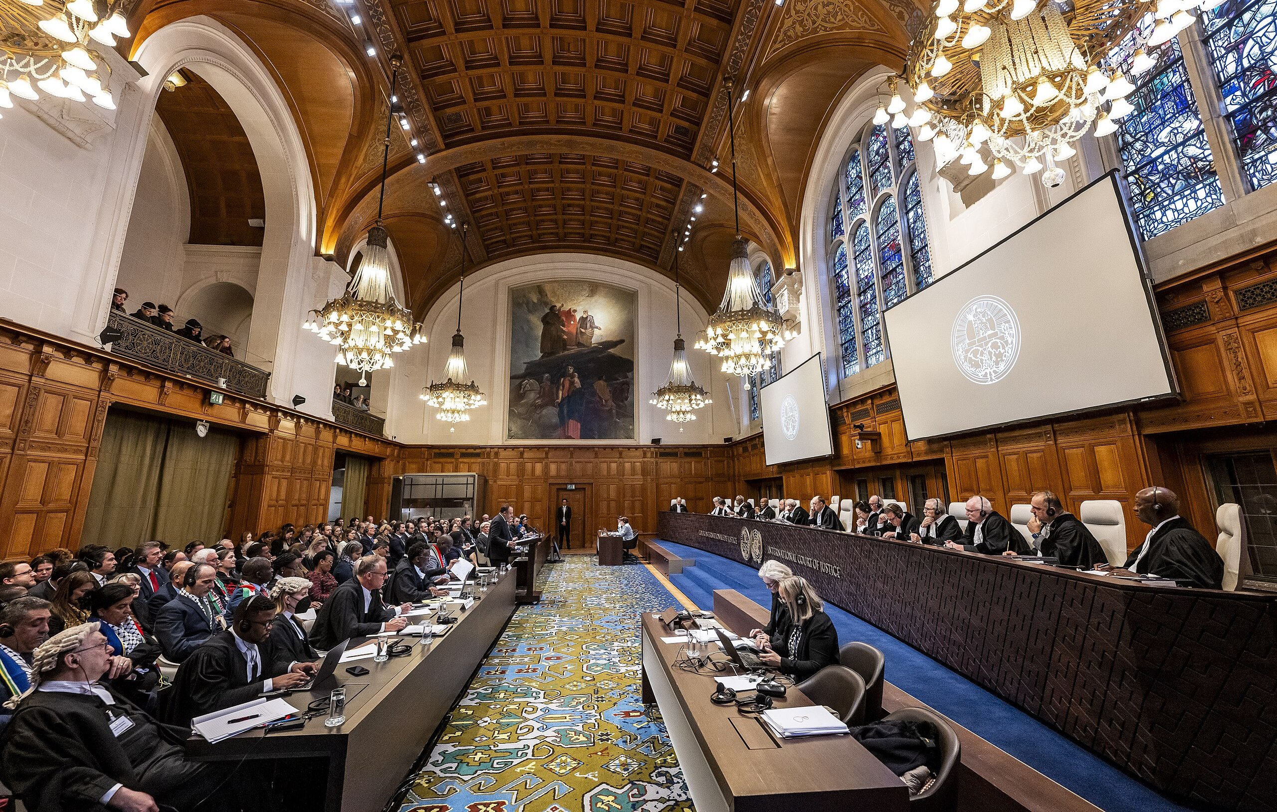The image depicts an interior of a grand courtroom, likely in an international court such as the International Court of Justice. The room features high wooden ceilings with intricate designs and ornate chandeliers providing warm lighting. Large windows with stained glass allow natural light to filter in. In the foreground, there are rows of judges seated on a raised platform, alongside legal representatives and observers. The layout includes a long table with officials on one side and a broad audience on the other. The design of the room is both formal and historic, emphasizing the importance of the proceedings taking place.