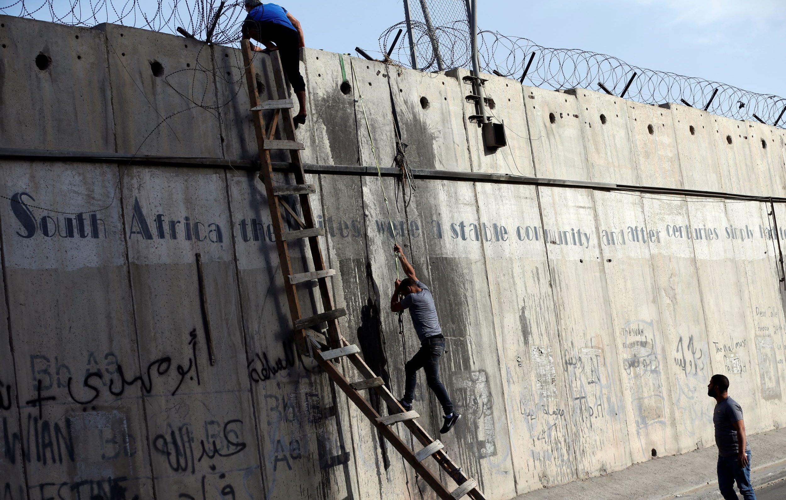 L'image montre une scène où deux personnes tentent de monter ou de descendre d'un mur de béton. Une échelle est appuyée contre le mur, et il y a des barbelés au sommet. Le mur présente également des graffitis ou des inscriptions, et l'environnement semble urbain. Les personnes semblent concentrées sur leur tâche, soulignant une dynamique d'effort et de défi.