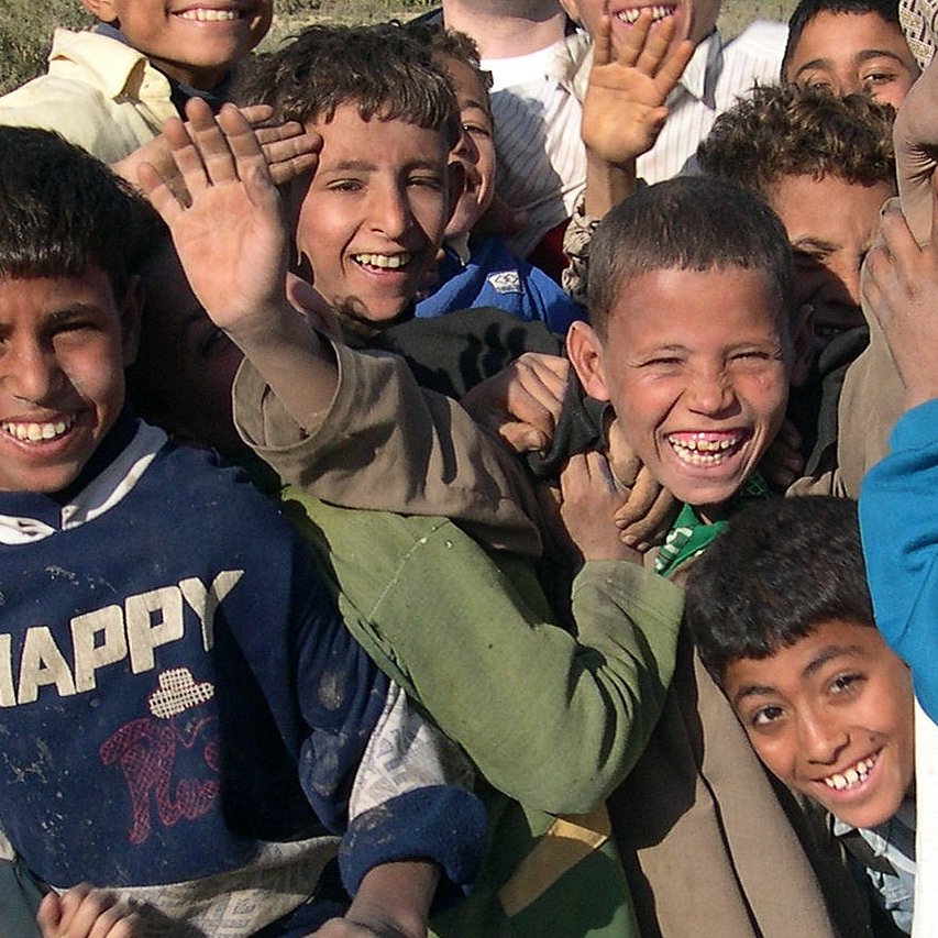 L'image montre un groupe d'enfants qui sourient joyeusement. Ils semblent apprécier le moment, avec des expressions de joie et beaucoup d'énergie. Les enfants sont vêtus de vêtements variés et certains lèvent la main ou se penchent vers l'avant, montrant un sentiment d'excitation et d'unité. L'arrière-plan est flou, mettant en avant leurs visages animés et heureux.