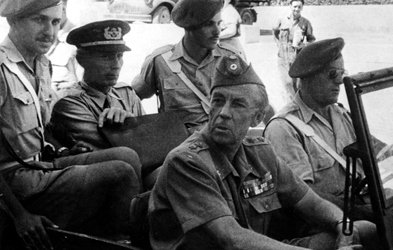 L'image montre un groupe de militaires assis dans un véhicule. Il y a plusieurs hommes en uniforme, portant des chapeaux distinctifs. Certains sont assis à l'avant, tandis que d'autres semblent être à l'arrière. L'atmosphère est solennelle, et il pourrait s'agir d'un moment lié à une opération ou une mission militaire. Le fond est flou, mais on aperçoit d'autres véhicules et un environnement urbain.
