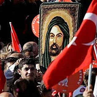 L'image montre une foule rassemblée lors d'une manifestation. Au centre, une personne tient une grande image représentant un personnage avec une longue barbe et un turban, entourée de drapeaux turcs. La foule semble engagée et énergique, avec des expressions variées sur les visages des participants.