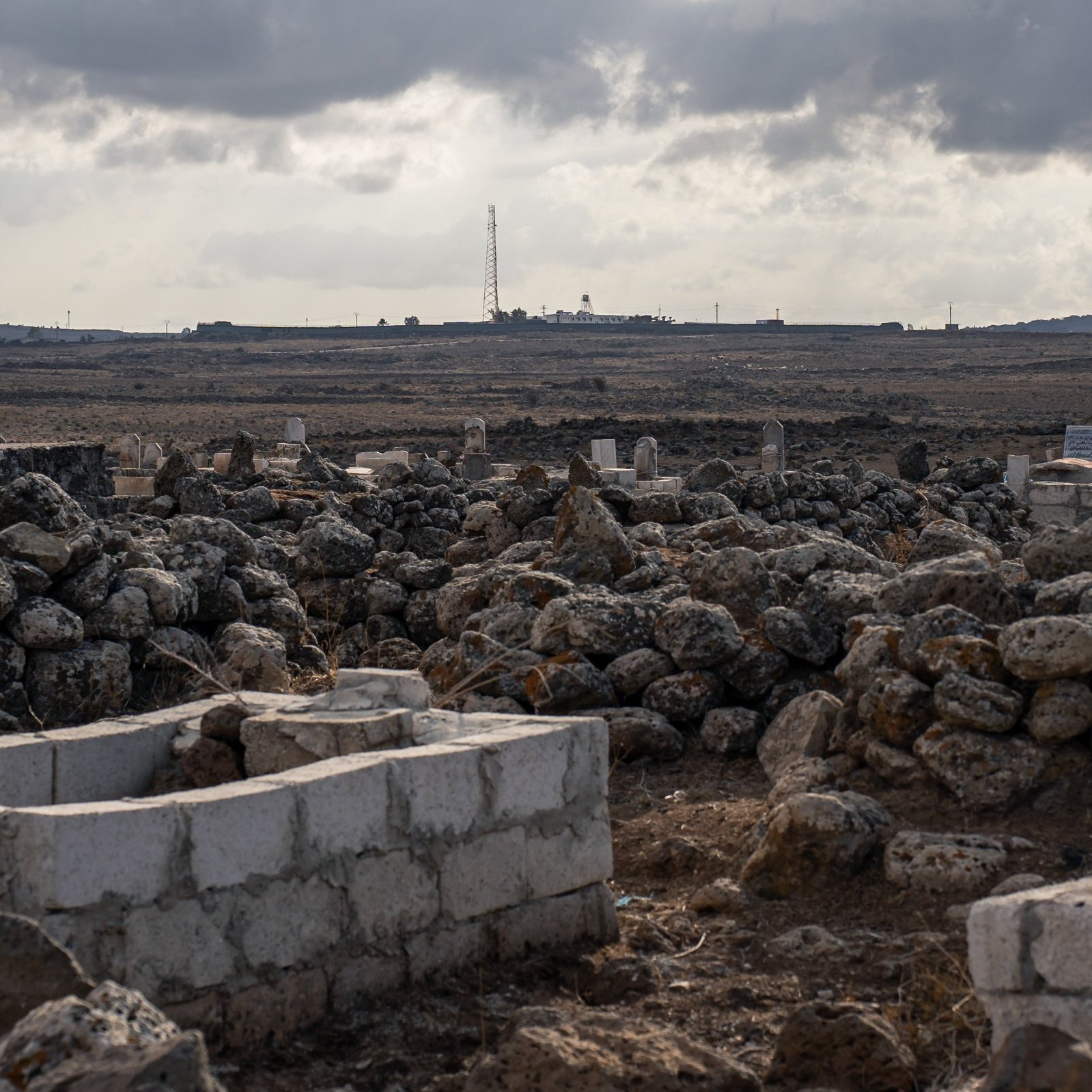 Un paisaje árido con tumbas y rocas, bajo un cielo nublado y distante, se ve una torre.