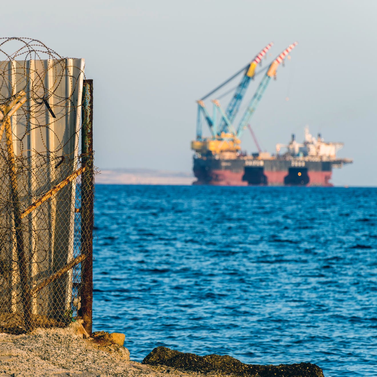 L'image montre une barrière en métal, probablement un mur de séparation, avec du fil barbelé à sa base. En arrière-plan, on aperçoit des barges ou des plateformes offshore, avec des grues, sur un fond d'eau calme. Le ciel semble dégagé, et l'ensemble donne une impression de contraste entre la structure humaine et l'environnement maritime.