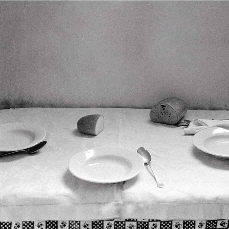 L'image montre une table couverte d'une nappe blanche, où se trouvent deux assiettes blanches, une cuillère et un couteau. Un petit pain est positionné sur un côté et un verre est renversé au centre de la table. L'ambiance générale est sobre et monochrome, ce qui donne une impression de simplicité et de calme. Les murs derrière la table sont légèrement ombragés, ajoutant un effet de profondeur à la composition.