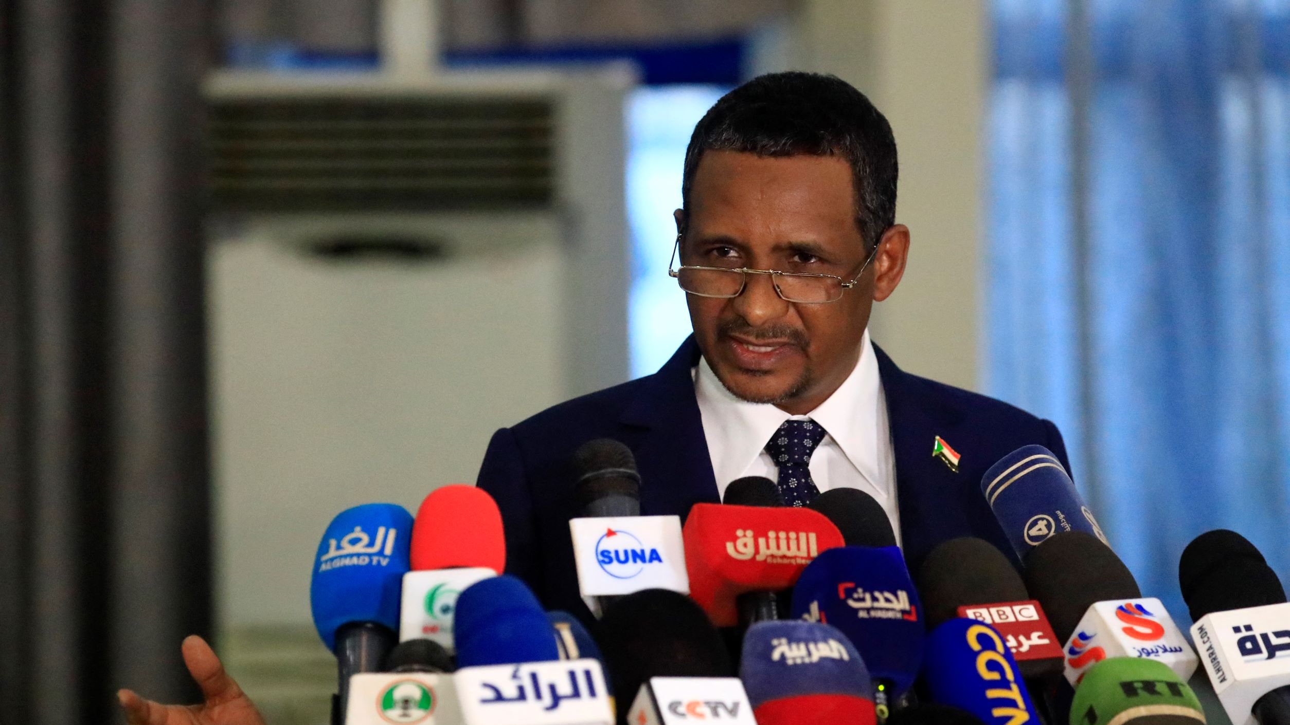 در این تصویر، مردی در حال صحبت کردن در جلوی میکروفن‌های زیادی است. او لباس رسمی به تن دارد و به نظر می‌رسد در حال ارائه یک سخنرانی یا بیانیه است. در پس‌زمینه، تعدادی دوربین و میکروفون وجود دارد که نشان‌دهنده جوی رسانه‌ای و توجه عمومی به صحبت‌های اوست. فضا به‌نظر رسمی و جدی می‌رسد.