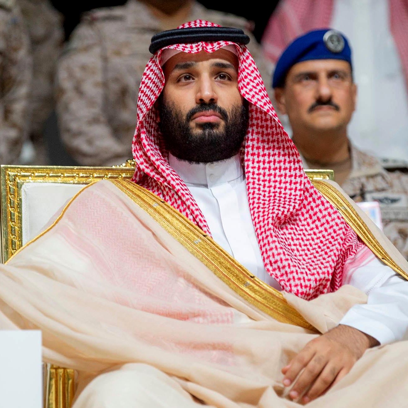 A man in traditional Saudi attire sits on a throne-like chair, looking serious and dignified.