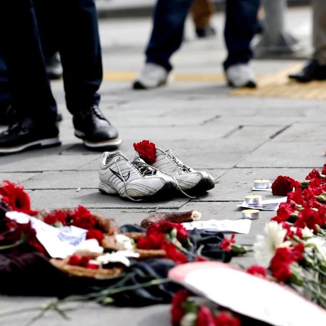 L'image montre un sol avec une paire de chaussures de sport blanches laissées à l'écart. Autour des chaussures, il y a une traînée de fleurs rouges, probablement des œillets, qui sont disposées en ligne. On peut également voir d'autres objets sur le sol, tels que des photos et des éléments décoratifs. En arrière-plan, des gens sont visibles, mais leur apparence et leurs détails ne sont pas clairement discernables. L'ensemble de la scène dégage une atmosphère de mémoire ou de commémoration.