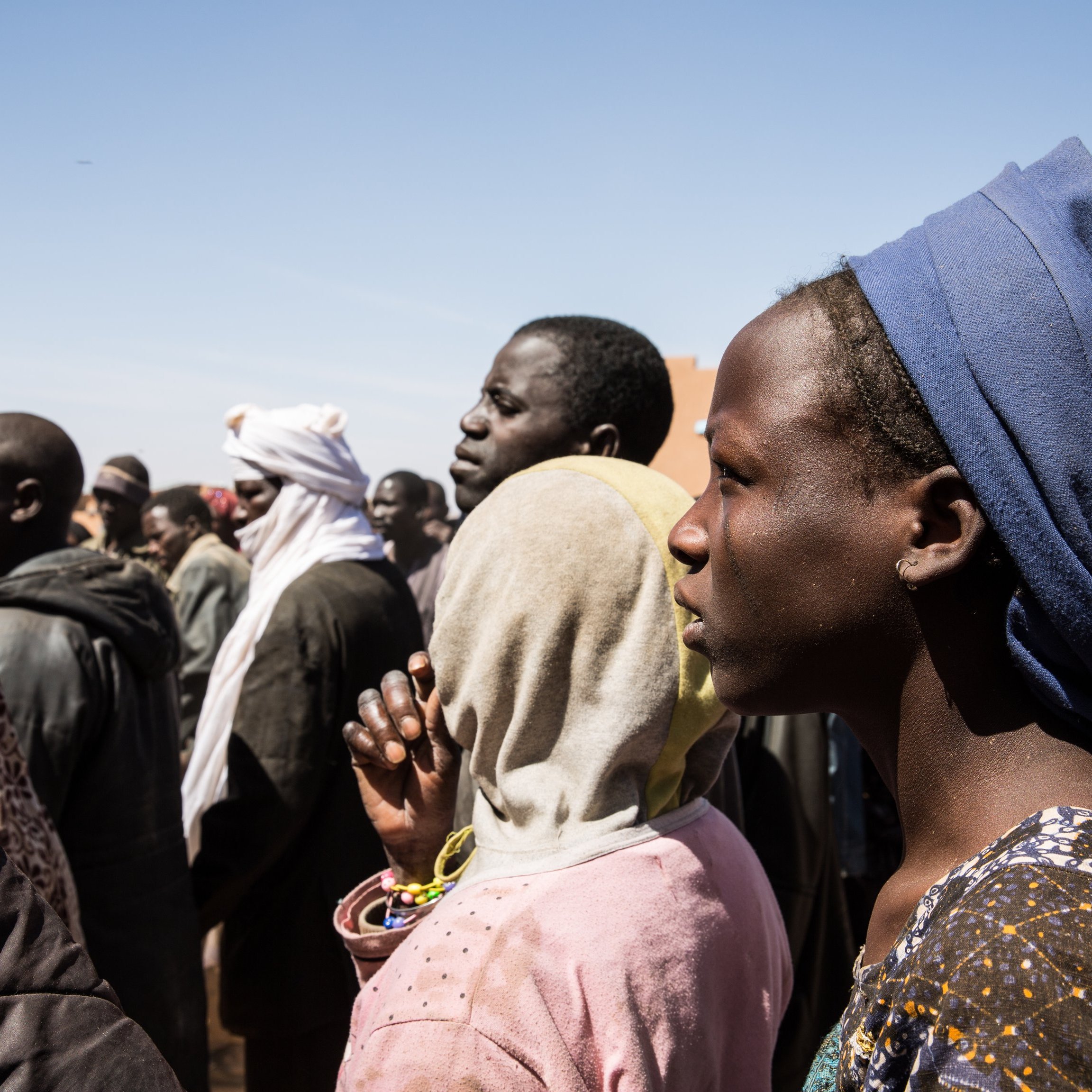 L'image montre un groupe de personnes, probablement en attente ou rassemblées pour une raison quelconque. On peut voir des visages divers avec des expressions sérieuses. Certaines personnes portent des vêtements traditionnels et des accessoires variés, comme des écharpes ou des foulards. L'arrière-plan est flou, mais le ciel est dégagé. L'ambiance semble concentrée, peut-être en lien avec un événement important.