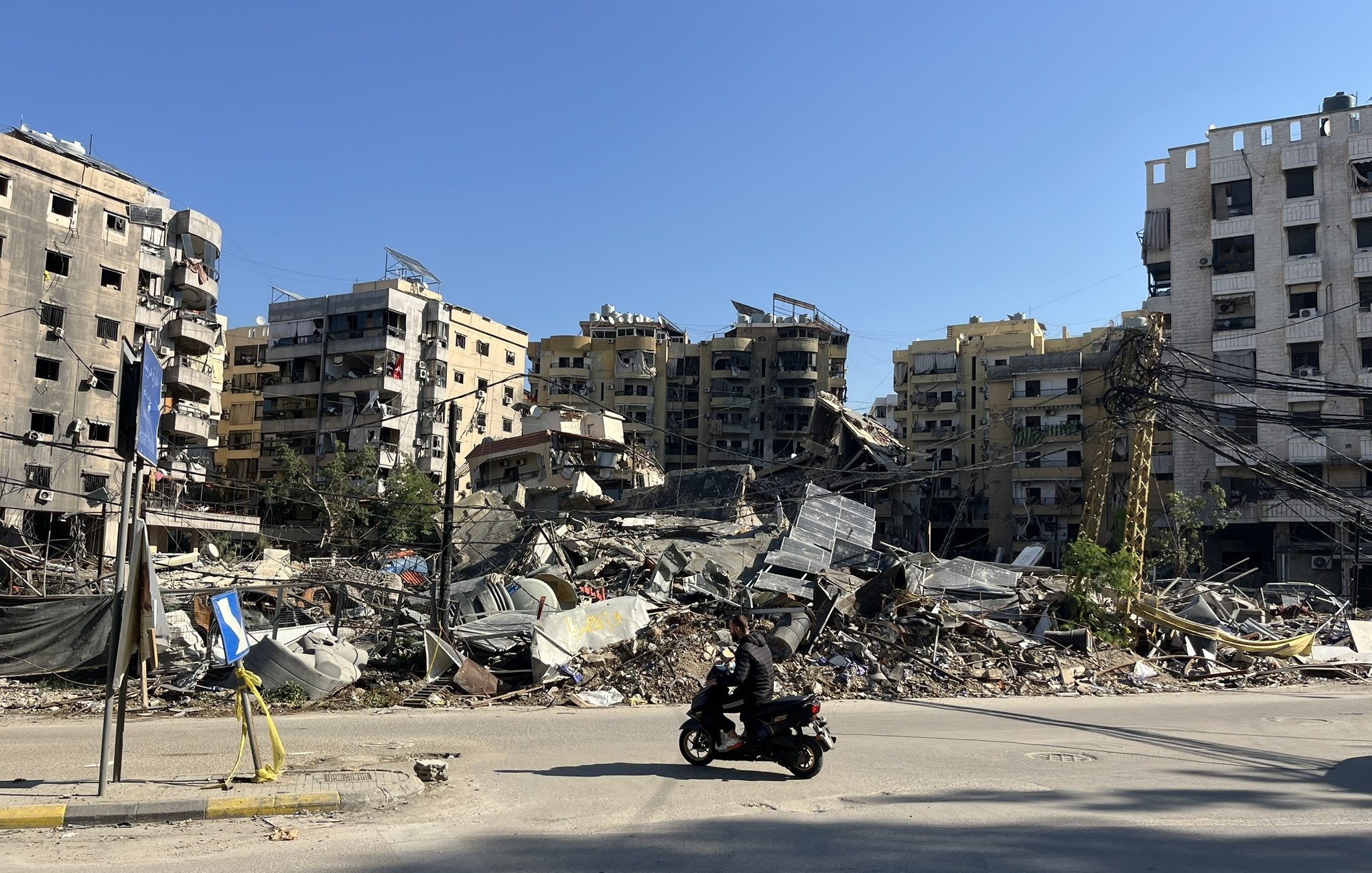L'image montre une scène de destruction urbaine, où plusieurs bâtiments sont partiellement effondrés et en ruines. On peut voir des décombres éparpillés au sol, tandis qu'une personne à moto passe devant. Le ciel est dégagé, laissant transparaître un jour ensoleillé, contrastant avec le désastre visible. La scène évoque un fort sentiment de désolation et de dégâts, typique de zones touchées par des conflits ou des catastrophes.