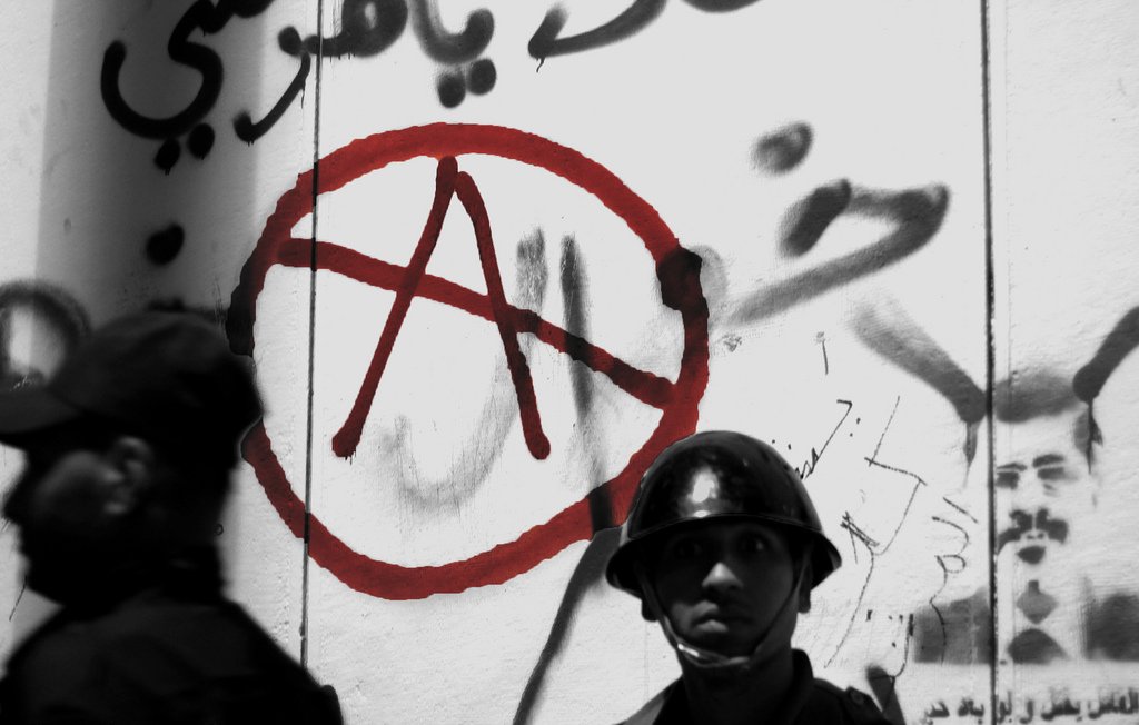 L'image présente un mur recouvert de graffitis, dont un symbole anarchiste stylisé en rouge, entouré d'un cercle. En arrière-plan, on voit deux hommes en uniforme, probablement des forces de l'ordre. L'un d'eux est au premier plan, portant un casque, avec une expression sérieuse. L'atmosphère de la photo suggère une tension sociale, accentuée par l'utilisation de couleurs sombres et le contraste avec le symbole rouge.