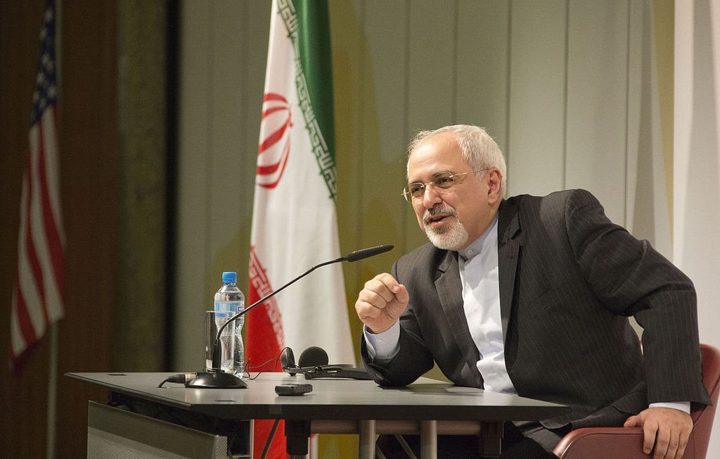 L'image montre un homme assis à une table de conférence, parlant avec animation. Il est habillé en costume, avec une chemise claire. À l'arrière-plan, on peut voir des drapeaux, y compris celui de l'Iran et celui des États-Unis. L'homme semble s'adresser à un public, probablement en répondant à des questions ou en faisant une déclaration importante.