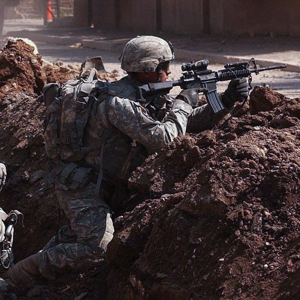 در این تصویر، دو سرباز در زمین‌های خاکی و سنگی جاگیر شده‌اند. آنها در حال استفاده از سلاح‌های خود هستند و به نظر می‌رسد در یک موقعیت جنگی یا تنش‌زا قرار دارند. پس‌زمینه تصویر به همراه فاکتورهایی مانند دود و خرابی، نشان‌دهنده یک محیط نظامی و چالش‌برانگیز است. لباس و تجهیزات سربازان نیز نشان‌دهنده آمادگی آنها برای عمل در شرایط سخت است.