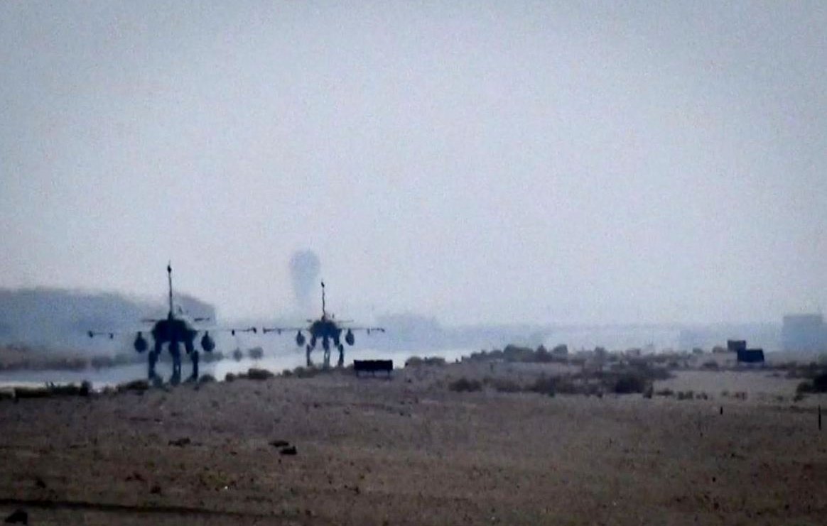 L'image montre deux avions militaires aperçus au loin dans un paysage désertique. L'atmosphère est légèrement brumeuse, ce qui donne une impression de profondeur. On peut distinguer une piste d'atterrissage ou d'envol, et le sol semble aride avec peu de végétation. Les avions sont visibles, mais les détails sont flous en raison de la distance et de la qualité de la prise de vue.