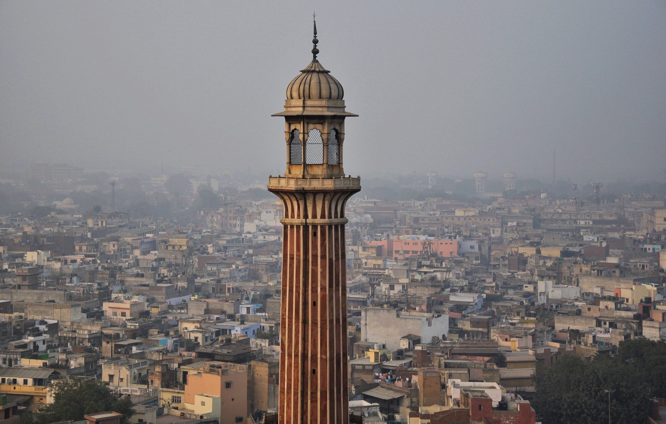 L'image montre un paysage urbain avec une tour élancée qui se dresse au milieu de nombreux bâtiments. La tour a une architecture distinctive, avec des motifs ornés et un dôme en haut. En arrière-plan, on peut voir une vaste étendue de maisons et de structures, témoignant d'une villeDense. L'atmosphère semble légèrement brumeuse, suggérant une pollution ou une humidité ambiante. Les couleurs des bâtiments varient, ajoutant une touche de diversité à l'ensemble de la scène.