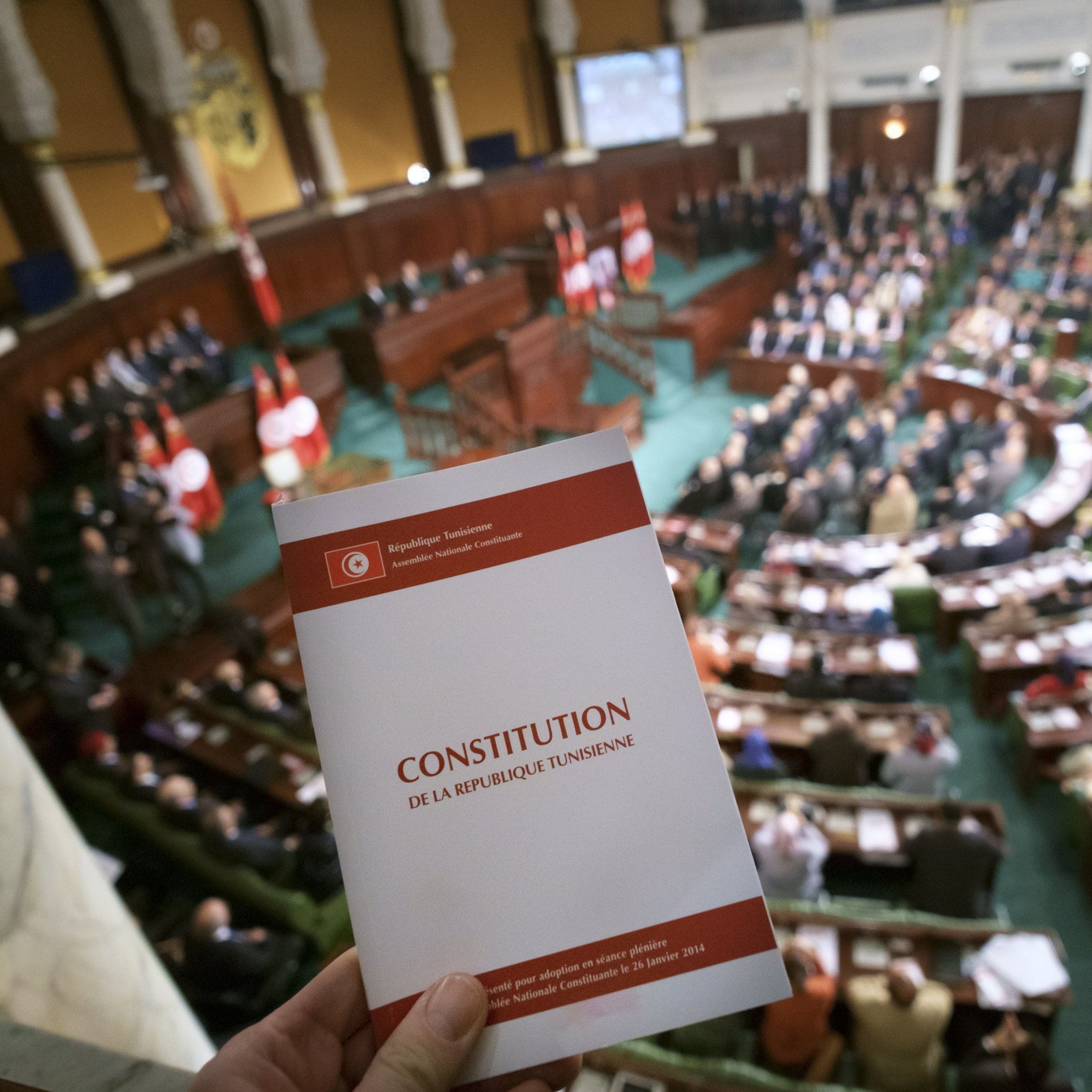 L'image montre une scène à l'intérieur d'une assemblée, probablement un parlement, avec un grand nombre de personnes assises dans une salle. Au premier plan, on voit une personne tenant un document portant le titre "CONSTITUTION DE LA RÉPUBLIQUE TUNISIENNE". L'assemblée est ornée de drapeaux, et l'ambiance semble solennelle, suggérant un moment important lié à la constitution ou à la législation.