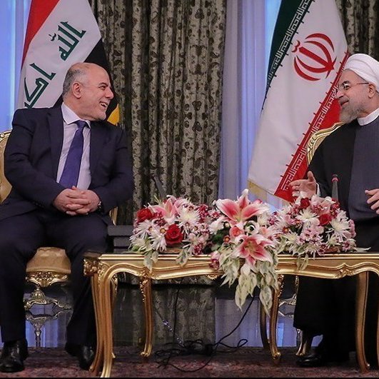 L'image montre une réunion entre deux hommes assis à une table décorée de fleurs. En arrière-plan, on peut voir les drapeaux de l'Irak et de l'Iran. Les deux personnages semblent en discussion amicale, avec des sourires et une ambiance conviviale. Le décor est élégant, avec des rideaux riches et un mobilier doré.