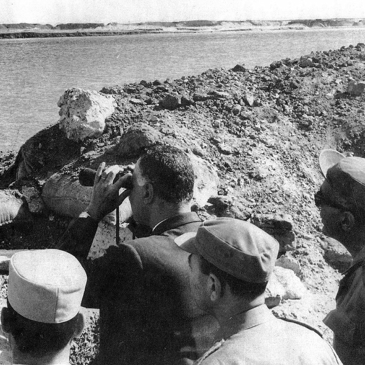 L'image montre un groupe d'hommes, dont l'un semble observer quelque chose au loin à l'aide de jumelles. Ils se tiennent près d'une rive d'eau, probablement à proximité d'un site de construction ou d'une digue, entourés de débris ou de terre. L'environnement semble suggérer une période historique, peut-être liée à des événements militaires ou politiques. Les hommes portent des uniformes ou des vêtements formels, ce qui indique qu'ils occupent probablement des positions officielles.