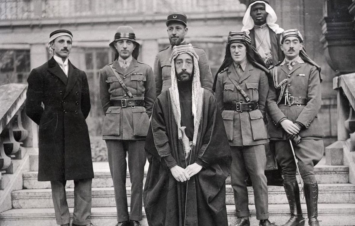 L'image montre un groupe de six hommes posant ensemble. Ils sont vêtus de différentes tenues militaires et traditionnelles. Au centre, un homme en robe traditionnelle, portant une tête couronnée, semble être le principal sujet. À sa gauche, un autre homme en uniforme militaire. Les autres sont également en uniformes variés, indiquant peut-être des origines et des rôles différents. L'arrière-plan montre une architecture qui semble historique. L'ensemble dégage une atmosphère de confluence culturelle et de collaboration.