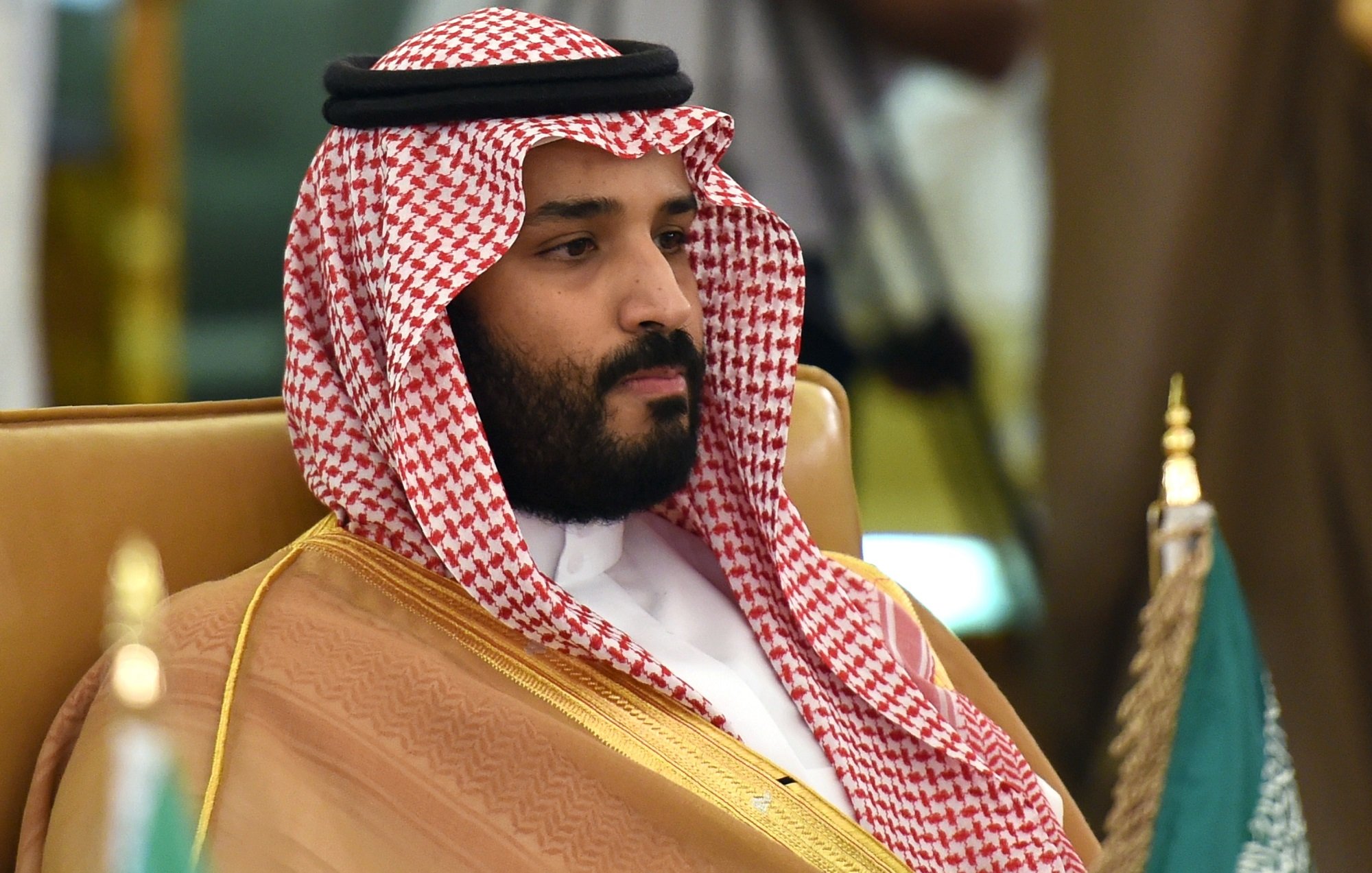 L'image montre un homme portant un long vêtement traditionnel arabe, composé d'une robe beige et d'un headdress appelé "ghutrah" ou "keffieh", qui est noué avec un bandeau noir. Il a une expression sérieuse et se trouve assis sur une chaise. En arrière-plan, des drapeaux peuvent être aperçus, suggérant un événement officiel ou une rencontre.