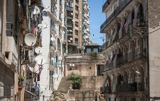L'image montre une vue urbaine d'une rue avec des bâtiments en pierre et des murs en béton. On observe un mélange d'architecture ancienne et moderne, avec des balcons ornés de fer forgé. De nombreux paraboles satellites sont installées sur les façades des bâtiments, tandis que des linges pendus à l'extérieur ajoutent une touche de vie quotidienne. Les escaliers menant à d'autres niveaux sont visibles, et le ciel est clair et ensoleillé, donnant une atmosphère vive à la scène.