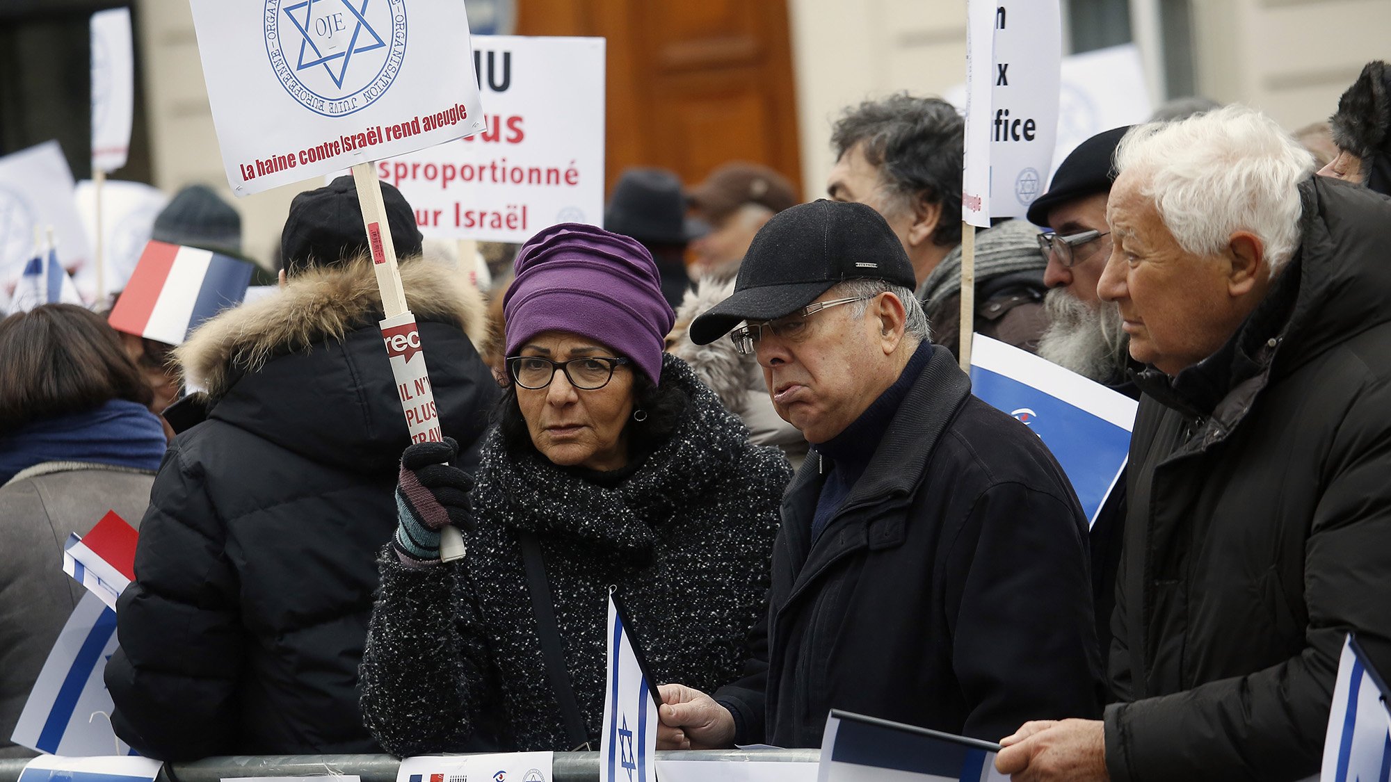 The image depicts a group of protesters gathered in an outdoor setting. They are holding signs and flags, including the Israeli flag and the French flag. The attendees appear to be engaged in a demonstration, showing solidarity or expressing opinions related to Israel. Some individuals are wearing winter clothing, suggesting it is during a colder season. The signs have various messages, possibly related to political or social issues concerning Israel. The atmosphere seems serious, highlighting a significant concern among the participants.