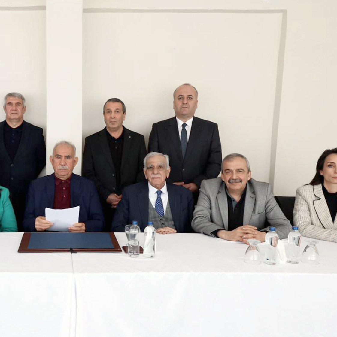 تصویر یک گروه از افراد را نشان می‌دهد که در یک جلسه رسمی نشسته‌اند. آنها به ترتیب در دو ردیف قرار دارند. میز روبروی آنها خالی از اشیا است و در پس‌زمینه، دیواری ساده و روشن وجود دارد. افراد لباس‌های رسمی پوشیده و به دوربین نگاه می‌کنند.