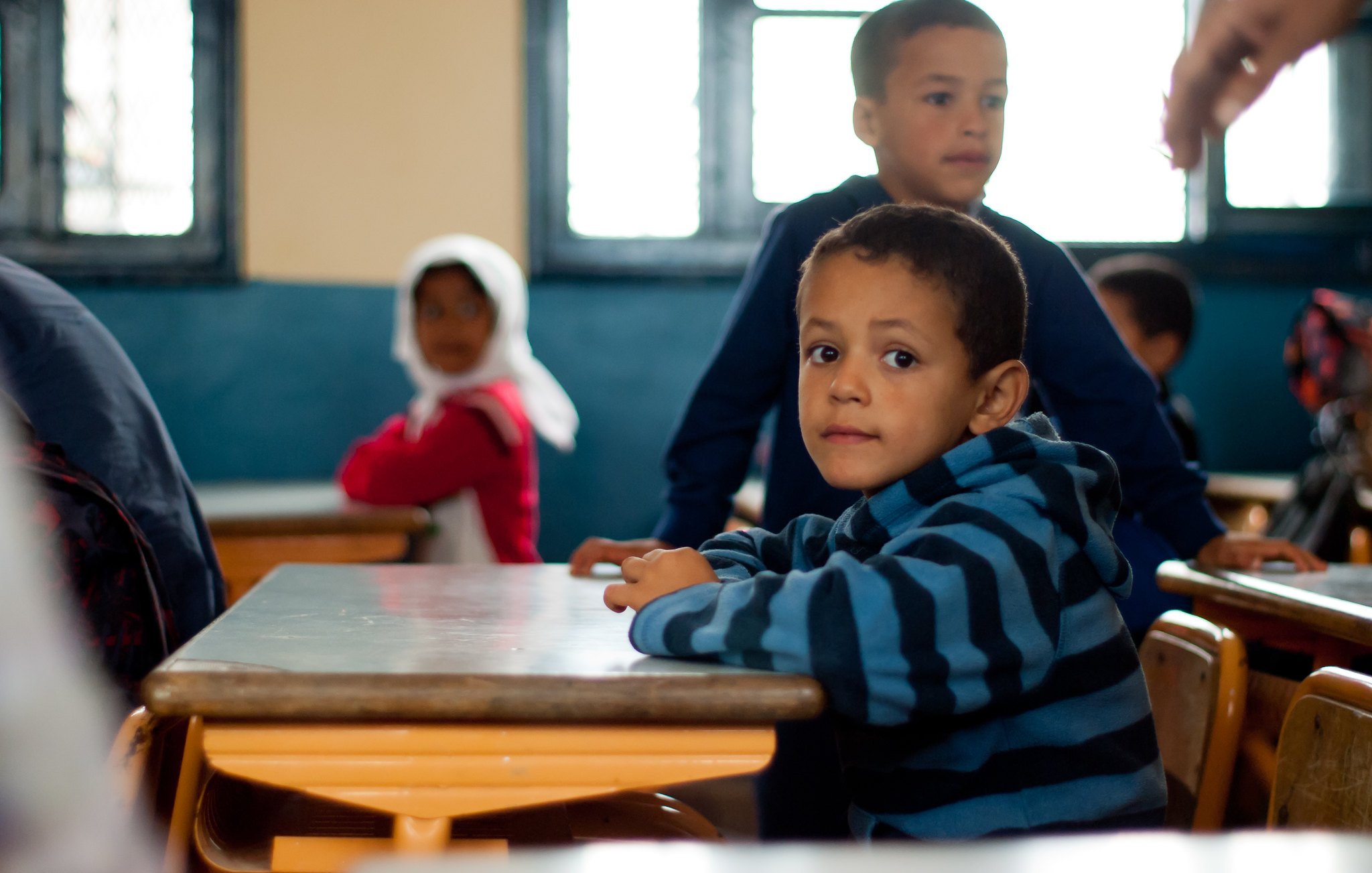 L'image montre une salle de classe où plusieurs enfants sont présents. Au premier plan, un jeune garçon avec un pull à rayures bleues et noires est assis à un bureau, regardant vers l'objectif avec une expression curieuse. À l'arrière-plan, on aperçoit d'autres enfants, dont un garçon debout et une fille avec un foulard. Les murs de la classe sont peints en bleu, et la lumière entre par des fenêtres. L'atmosphère semble calme et concentrée.