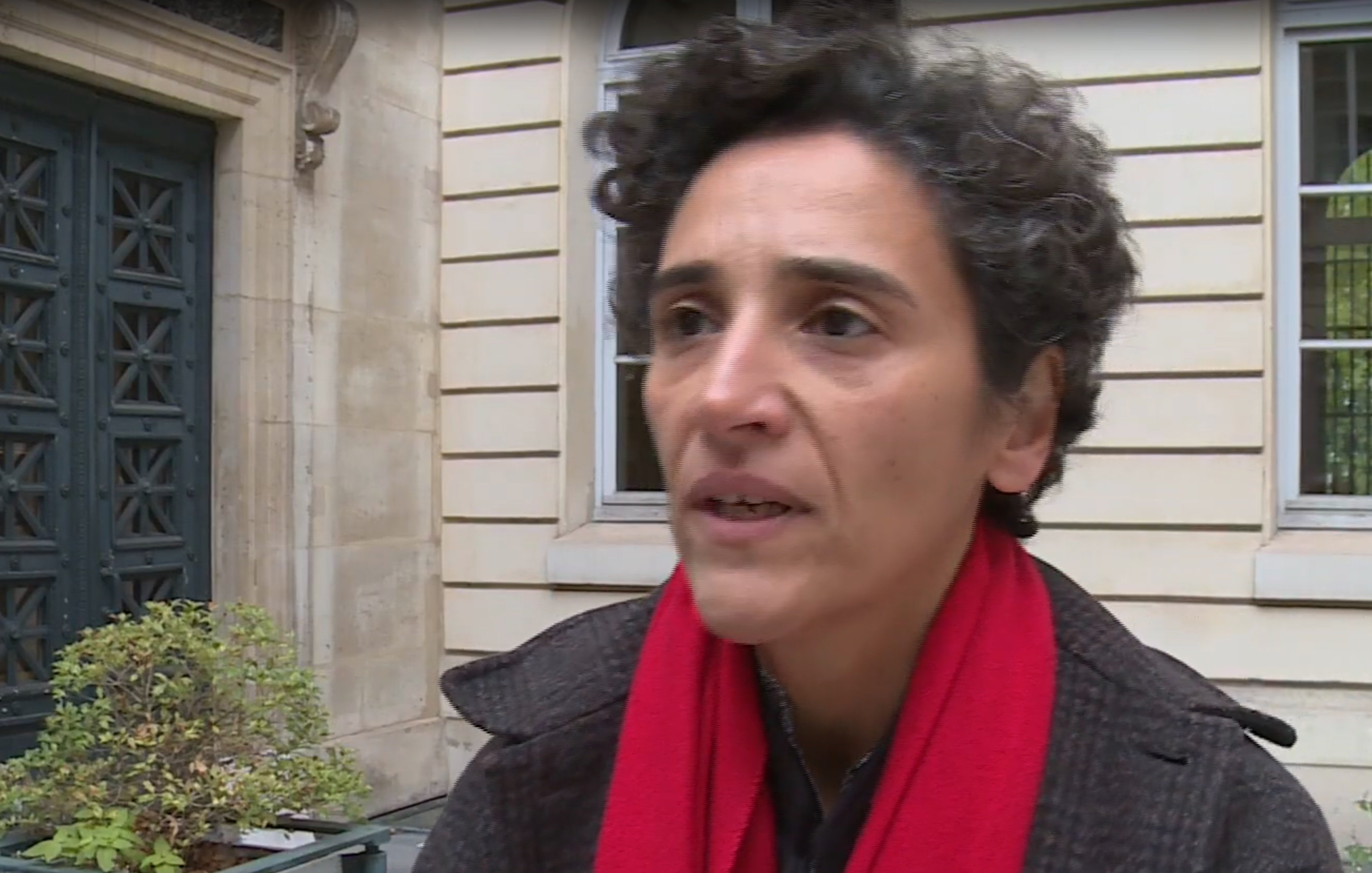 L'image montre une femme avec des cheveux bouclés, portant un manteau sombre et une écharpe rouge. Elle semble s'exprimer avec sérieux, peut-être dans le cadre d'une interview. En arrière-plan, on aperçoit un bâtiment avec des fenêtres et une porte d'entrée, ce qui donne une impression urbaine et architecturale.