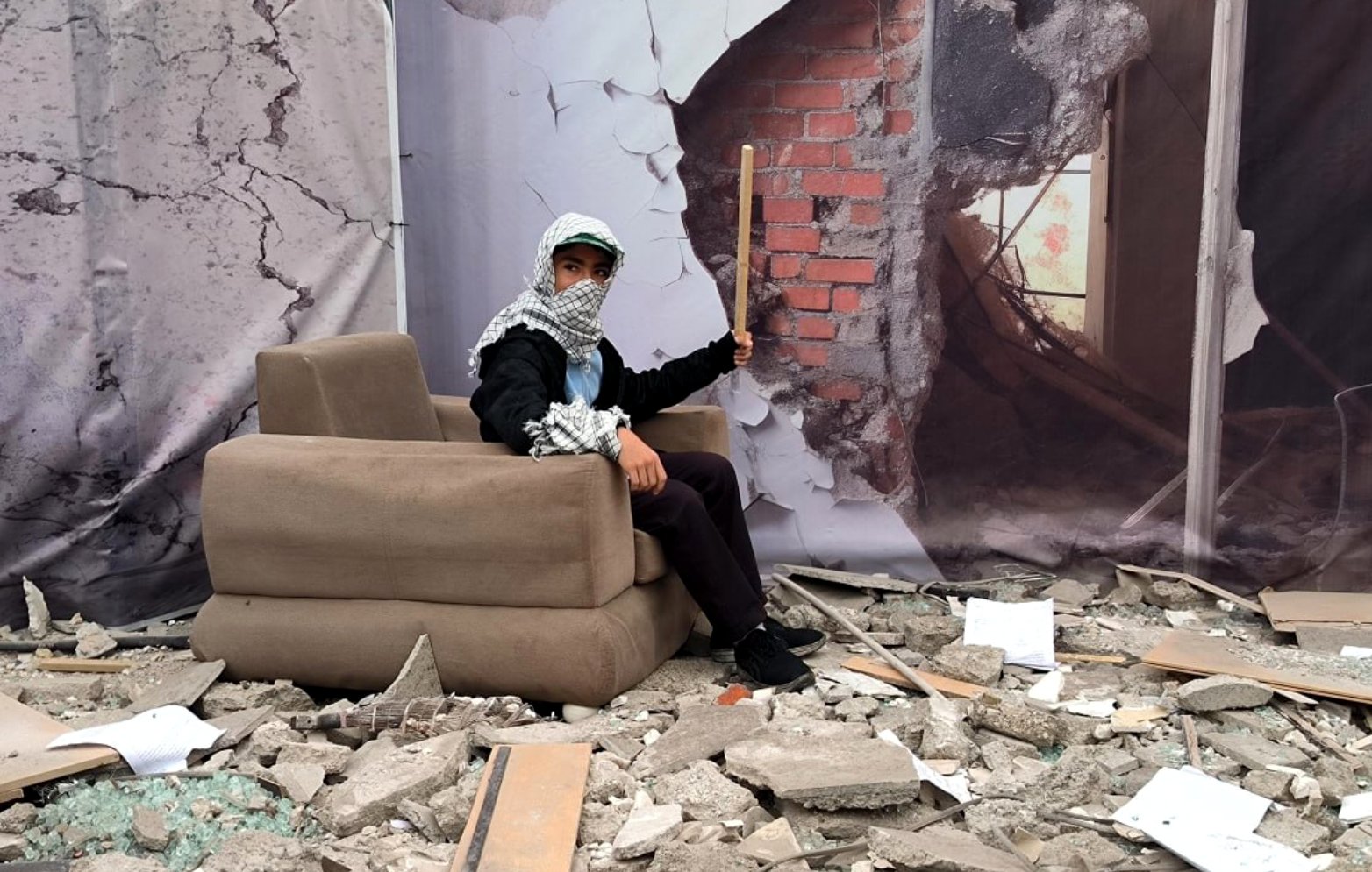 L'image montre une scène de destruction avec des débris éparpillés au sol, tels que des morceaux de béton et des morceaux de verre brisé. Au centre, une personne est assise sur un canapé apparemment plus ou moins intact. Elle porte un foulard autour de la tête et tient un bâton. En arrière-plan, il y a une illustration d'un mur brisé, ajoutant à l'impression de désolation et de chaos de la scène.