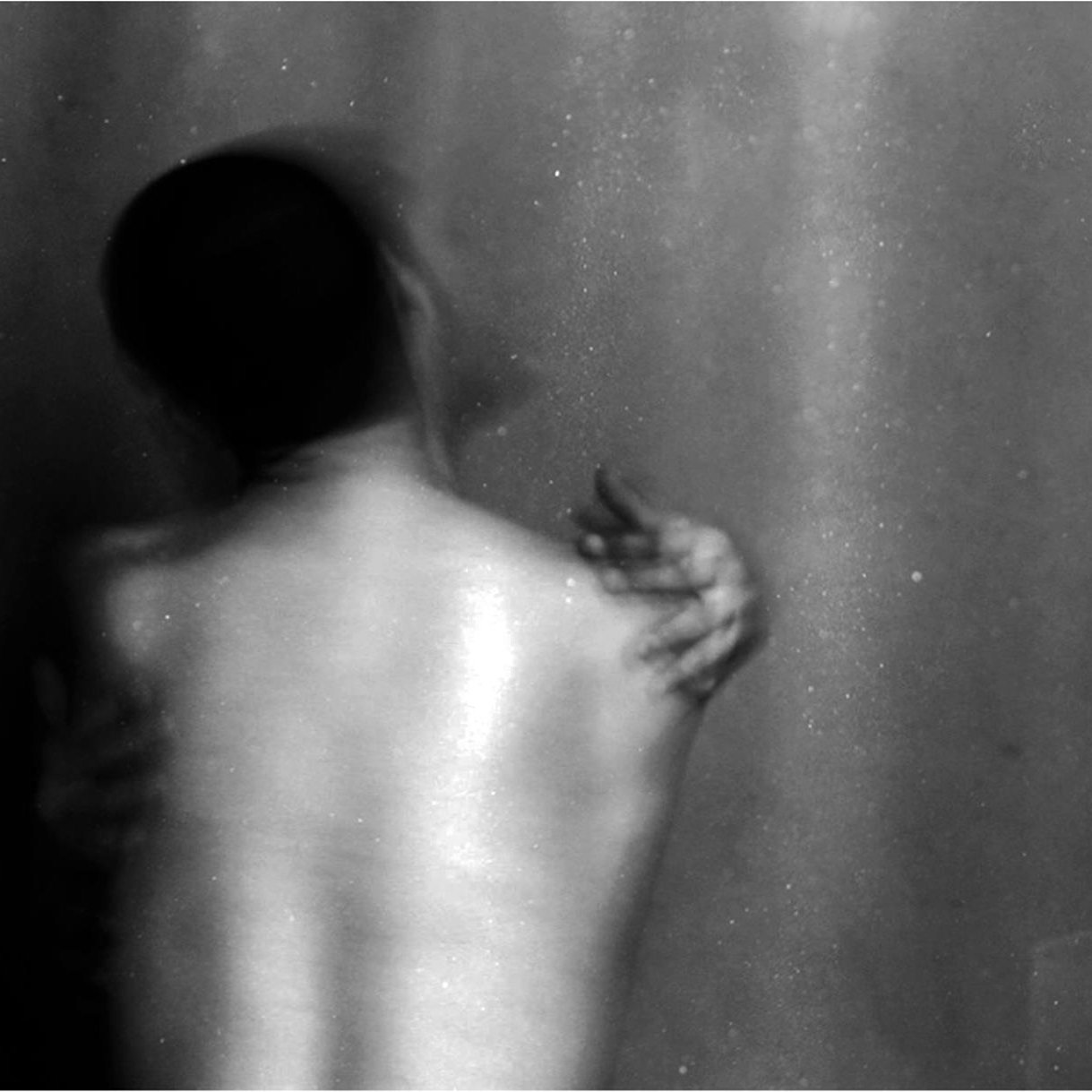L'image montre une silhouette floue d'une personne de dos, avec un éclairage doux qui fait ressortir les contours de son corps. L'arrière-plan est sombre, créant une atmosphère intime et mystérieuse. La posture de la personne évoque une certaine vulnérabilité, tandis qu'une main semble s'accrocher à son épaule, suggérant une interaction ou un lien émotionnel. L'utilisation du noir et blanc renforce l'aspect dramatique et introspectif de la scène.