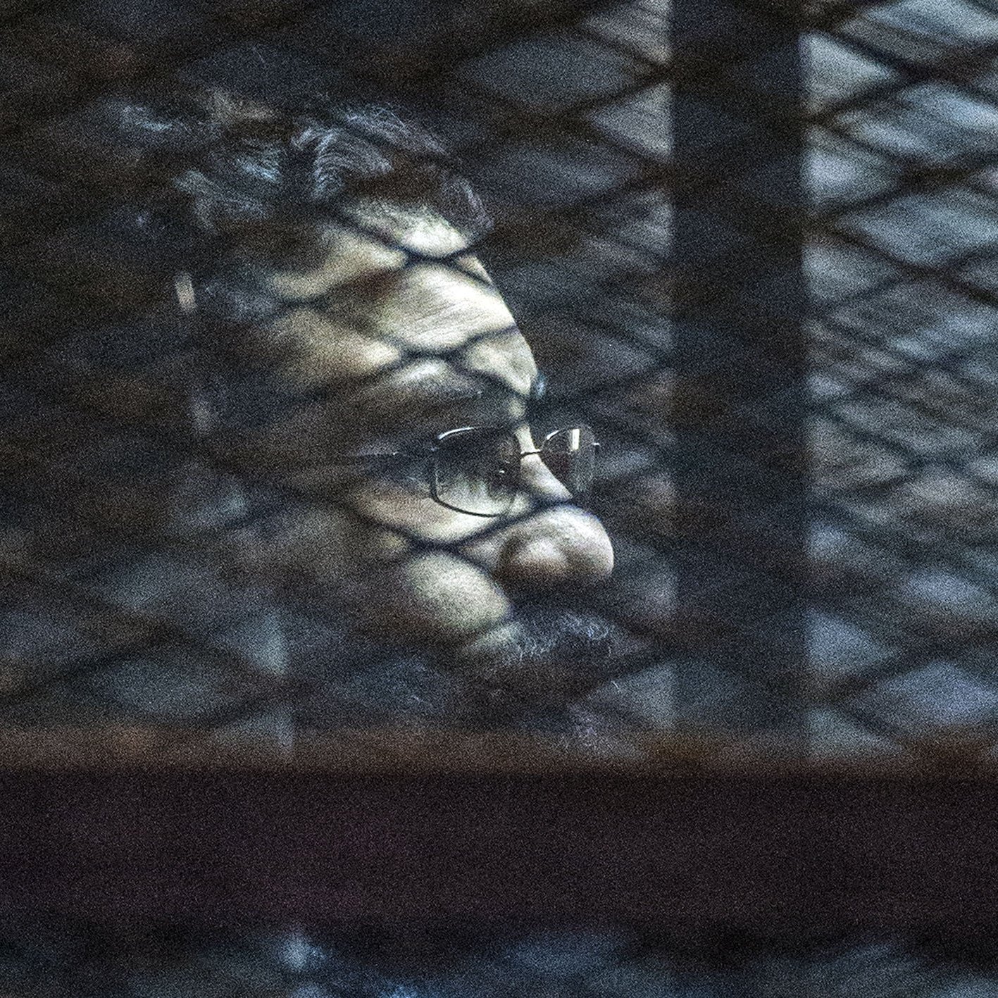 L'image montre un homme avec des lunettes et une barbe, dont le visage est partiellement obscurci par des ombres. On aperçoit des mailles ou des barres qui créent un effet de grille à l'avant, suggérant qu'il se trouve derrière une sorte de barrière ou de grillage. L'éclairage de la scène est sombre, contribuant à une ambiance mystérieuse.