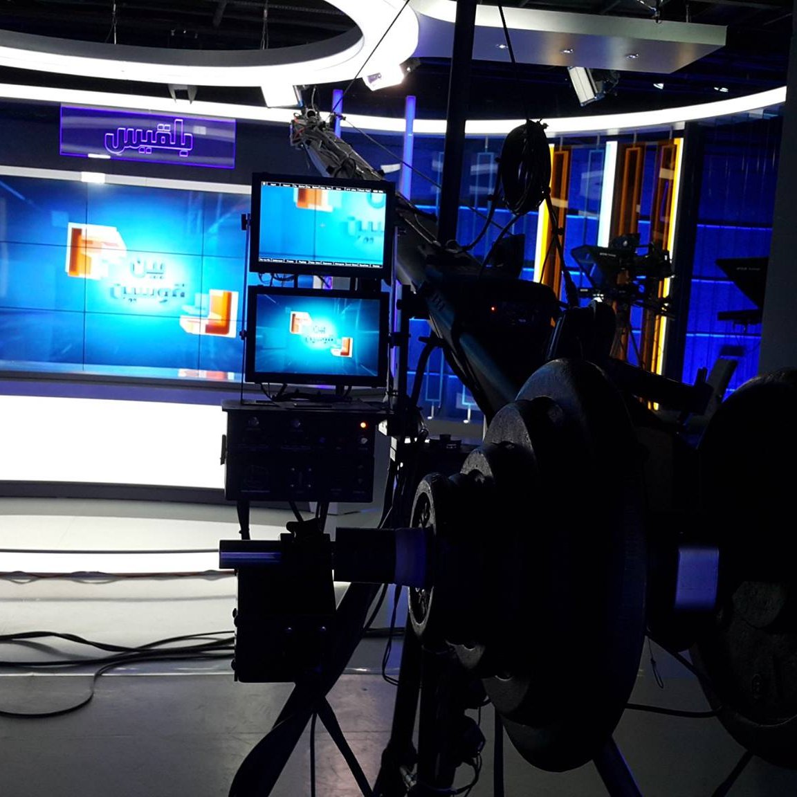 L'image montre un studio de télévision moderne. On peut y voir des lumières bleues qui éclairent l'espace, avec un fond décoratif et des écrans de présentation. À l'avant-plan, il y a une caméra sur un trépied, ainsi que des équipements techniques, ce qui indique que le studio est préparé pour une émission en direct ou un enregistrement. L'ambiance semble professionnelle et bien équipée pour la diffusion.