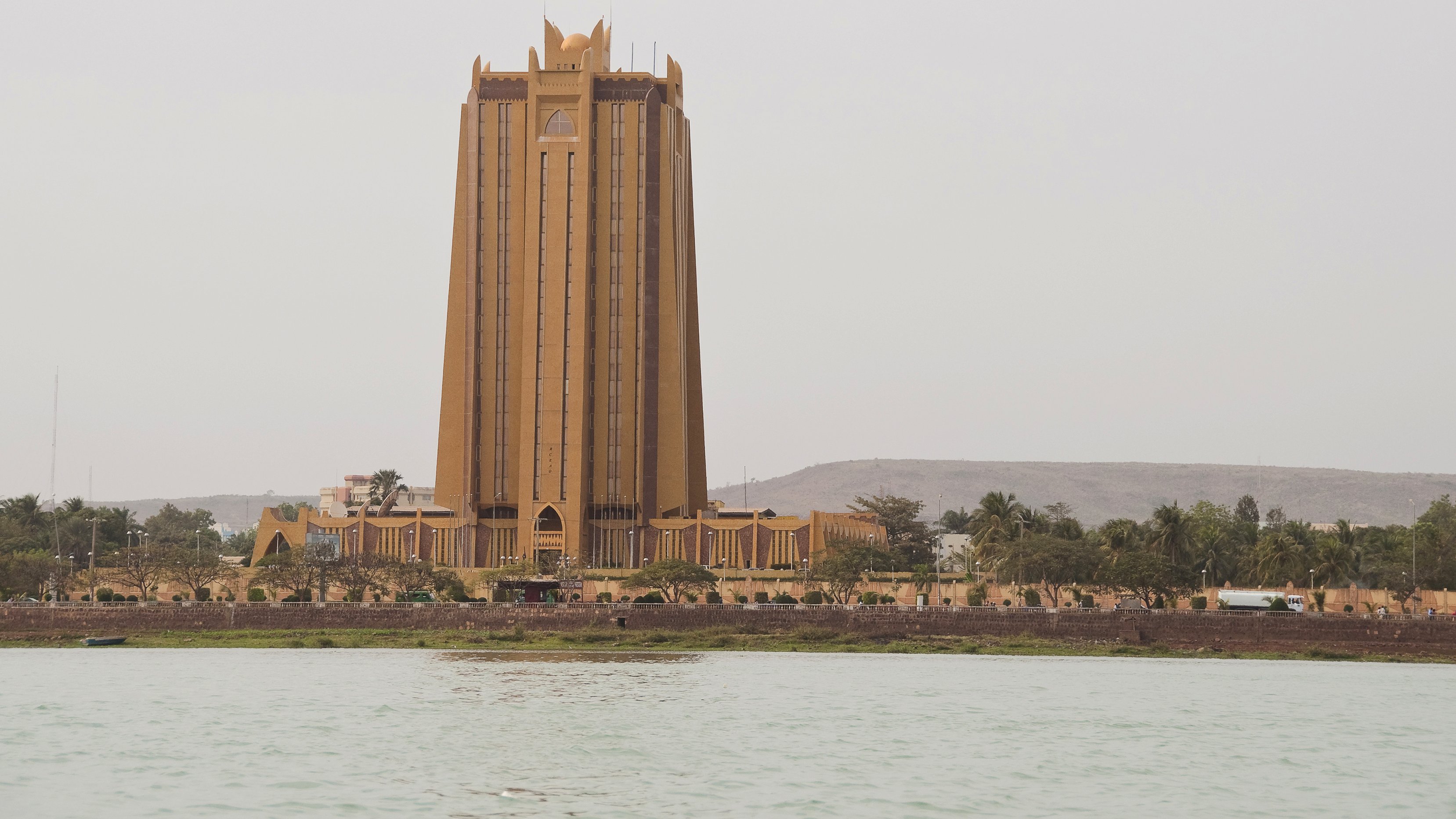 این تصویر نمای یک ساختمان بلند و برجسته را نشان می‌دهد که به رنگ زرد مایل به قهوه‌ای است. ساختمان دارای طراحی خاص و مدرنی است و با چندین طبقه و نوک‌های تیز در بالای آن مشخص می‌شود. در پیش‌زمینه، آب وجود دارد که به زیبایی تصویر می‌افزاید، و در پس‌زمینه، تپه‌ها و نواحی سبز دیده می‌شود. فضای عمومی اطراف ساختمان نیز مشخص است.