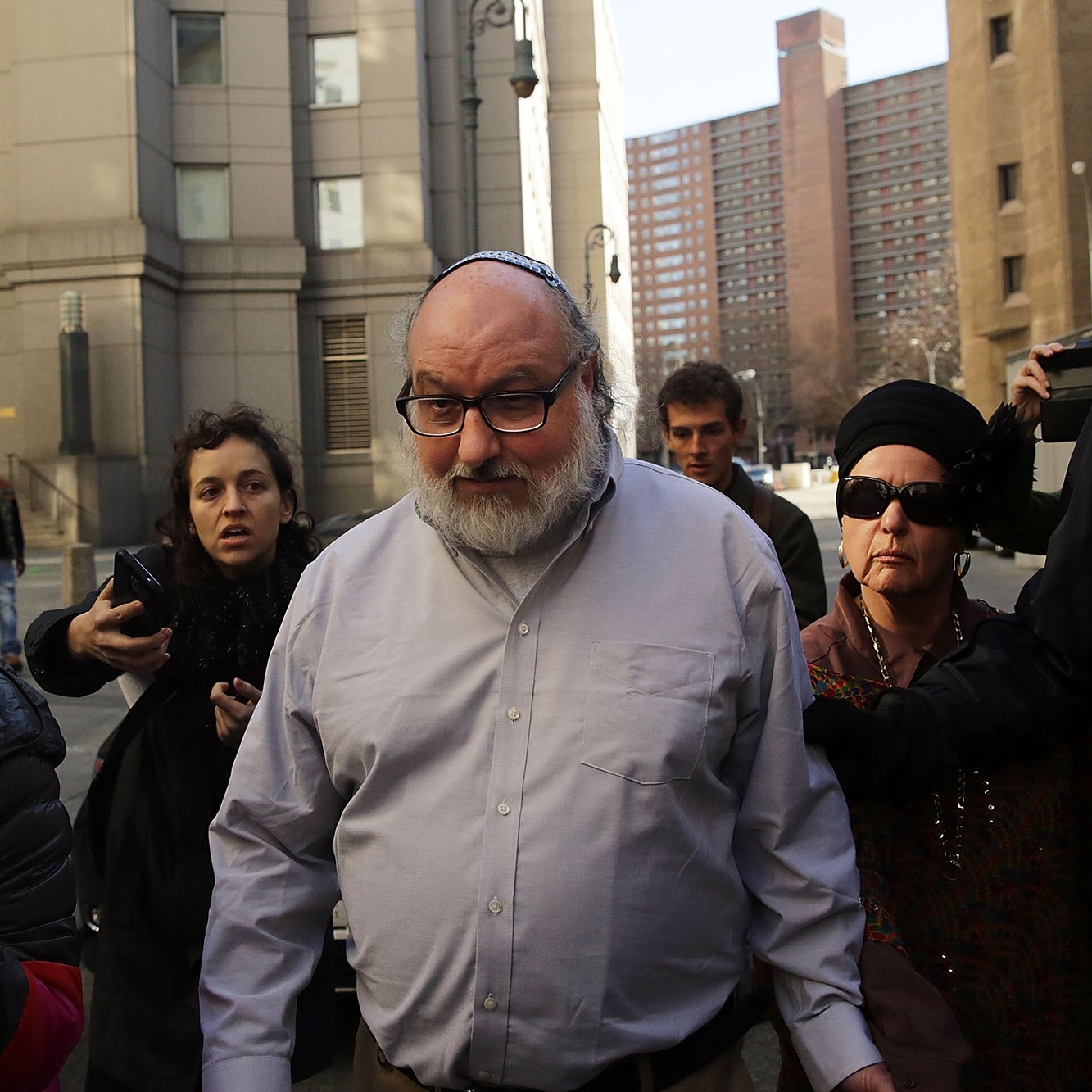 در این تصویر، یک مرد میانسال با عینک و کلاه کیپا در حال خروج از یک ساختمان به نظر می‌رسد. او به سمت دوربین‌ها و گروهی از خبرنگاران توجه دارد. چند زن هم در اطراف او هستند که یکی از آن‌ها عینک دودی به چشم دارد و دیگری کلاهی بر سر دارد. محیط اطراف شهری به نظر می‌رسد و چندین ساختمان بلند در پس‌زمینه قابل مشاهده است. جو تصویر حس هیجان و توجه رسانه‌ها را منتقل می‌کند.