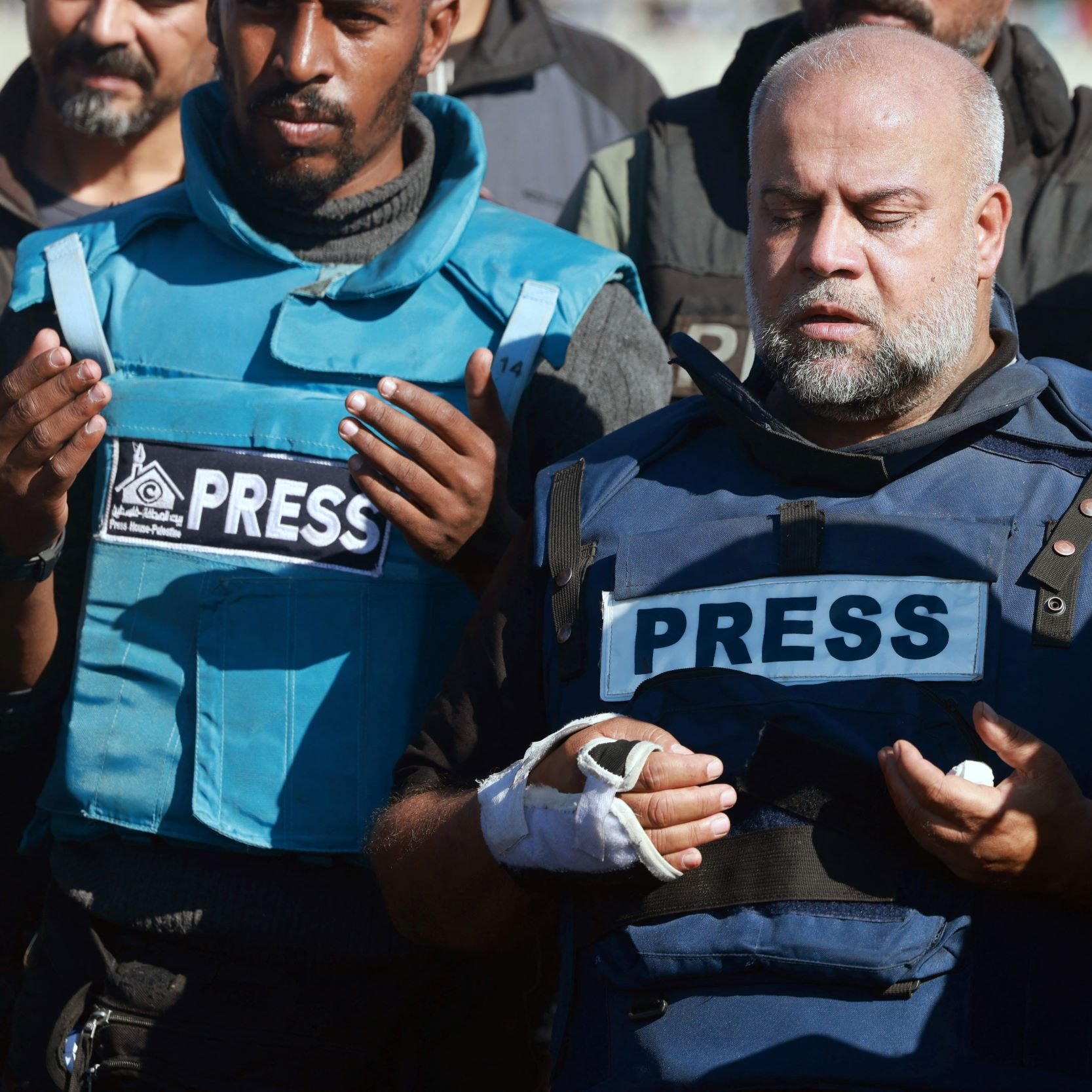 تظهر الصورة مجموعة من الأشخاص يرتدون سترات تحمل علامة "PRESS". يبدو أن أحدهم، الذي يرتدي سترة زرقاء، يتوسطهم ويبدو وكأنه في حالة تفكير أو دعاء، حيث يرفع يديه للأعلى. يبدو على وجهه وقور وجاد. باقي الأشخاص يظهرون وهم يشاركونه في هذه اللحظة، مما يعكس أجواء من التأمل أو التضرع.背景背景背景背景