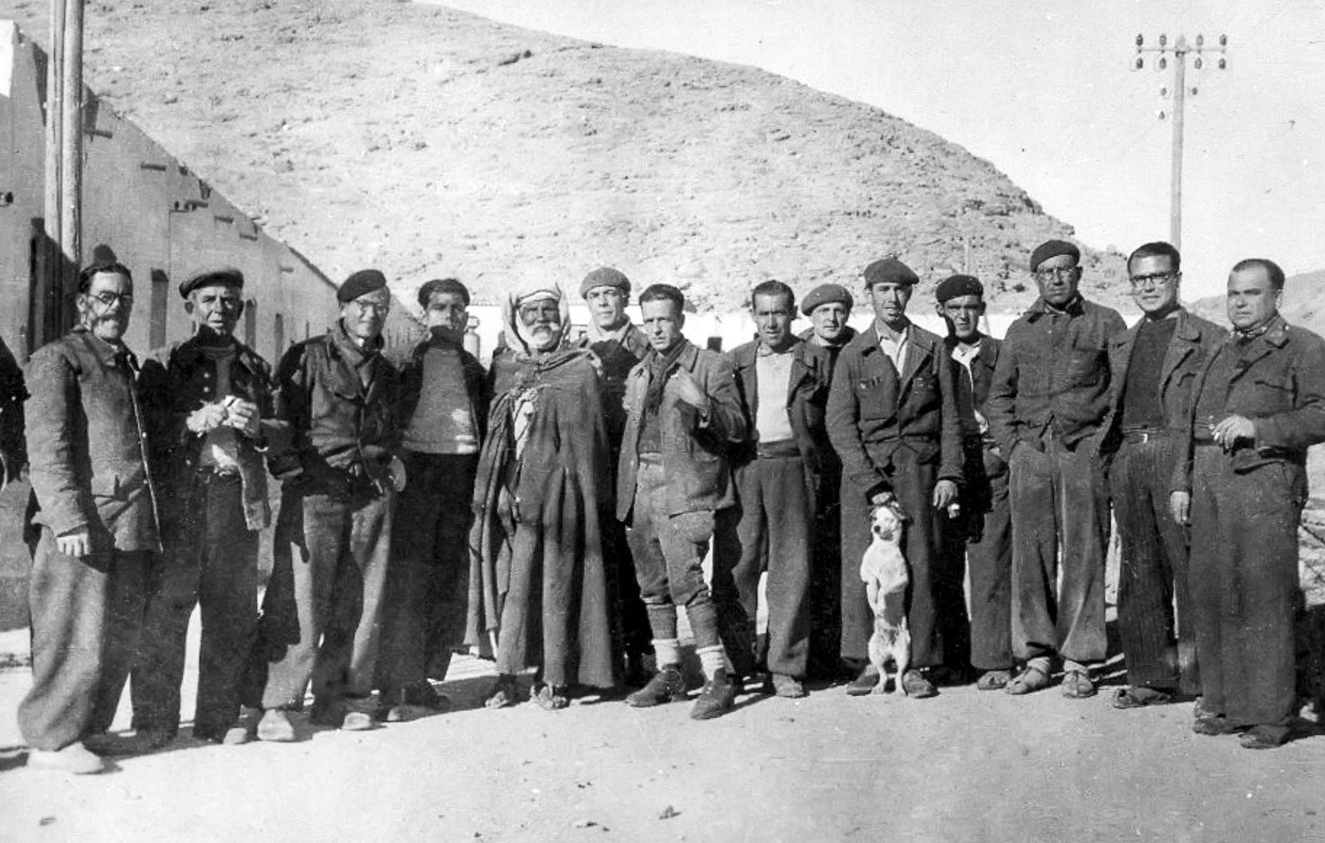 در این تصویر گروهی از مردان در یک محیط بیرونی تجمع کرده‌اند. آن‌ها به طور رسمی لباس پوشیده‌اند و در کنار هم ایستاده‌اند. در میان آن‌ها یک مرد با لباس سنتی و یک سگ دیده می‌شود. پس‌زمینه تصویر شامل تپه‌های خشک و ساختمان‌هایی به نظر می‌رسد که ویژگی‌های یک منطقه کوهستانی را نشان می‌دهد. نور تصویر قدیمی به نظر می‌رسد و به حالت سیاه و سفید است که به اثر حس نوستالژی خاصی می‌بخشد.