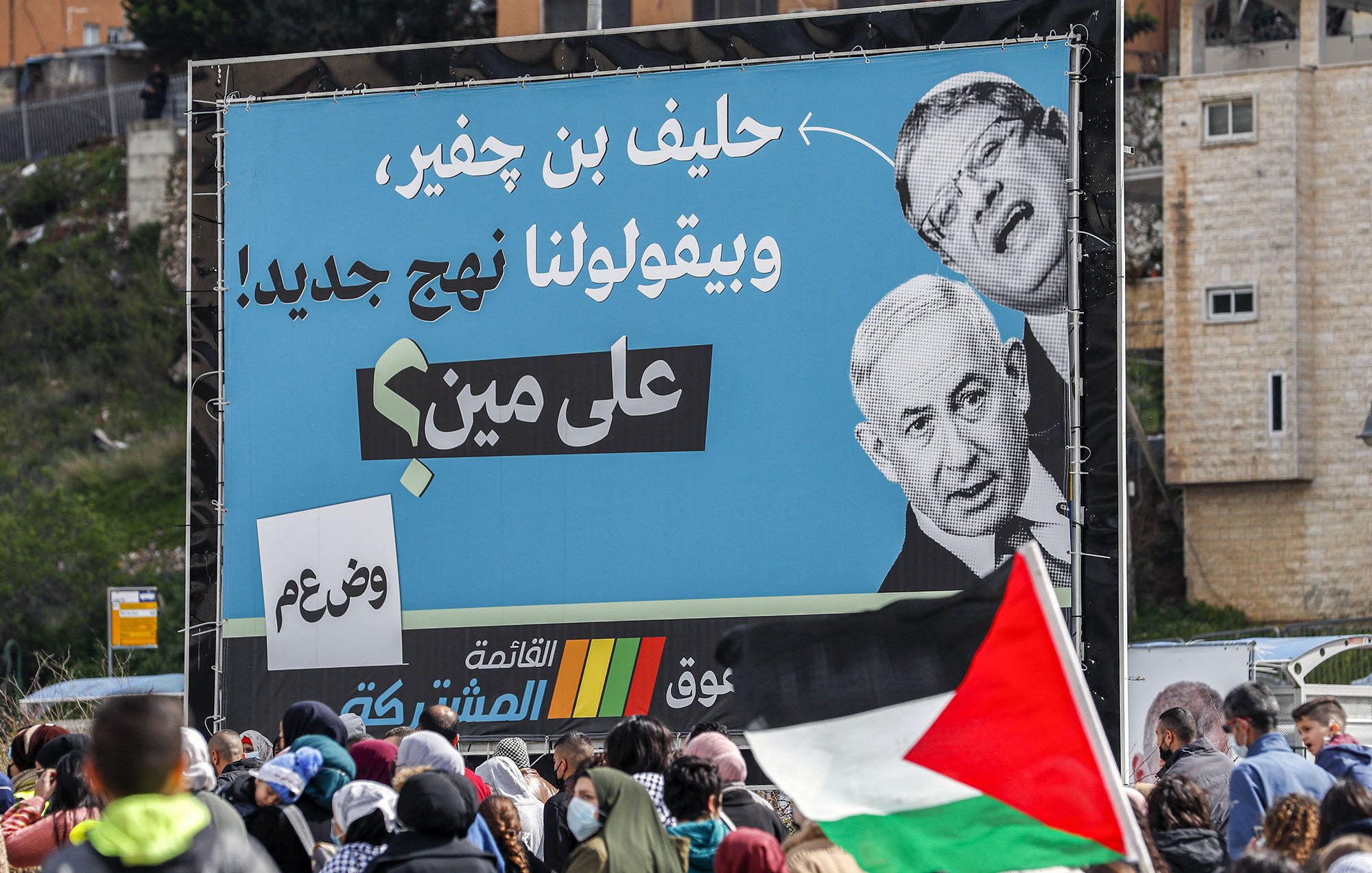 L'image montre une grande affiche sur un panneau publicitaire, présentant des portraits de deux hommes, dont l'un semble être un homme politique conversationnel. En arrière-plan, on peut voir une foule de personnes, certaines tenant des drapeaux palestiniens. L'affiche semble véhiculer un message politique, avec des slogans en arabe, suggérant un contexte de mobilisation ou de protestation. L'ambiance est dynamique, reflétant une situation de rassemblement public.