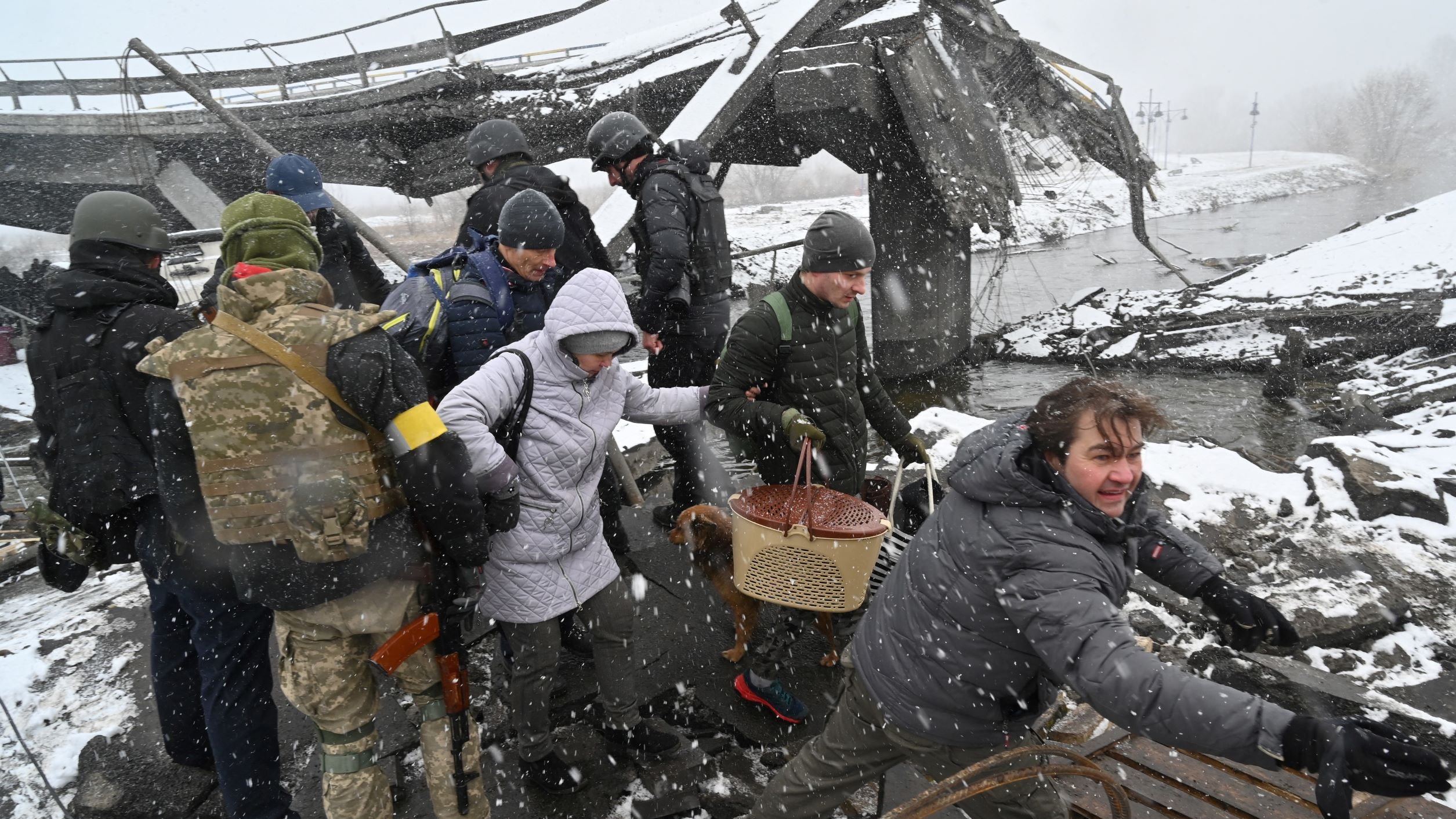 L'immagine mostra un gruppo di persone in un'area distrutta, probabilmente a causa di un conflitto o di un disastro. Alcune persone indossano abbigliamento militare, mentre altre sembrano civili. È in corso un salvataggio o un'evacuazione, con qualcuno che sta aiutando un'altra persona a attraversare il terreno difficile. La scena è caratterizzata da neve e condizioni atmosferiche difficili, suggerendo un ambiente critico e di emergenza. I volti mostrano preoccupazione e determinazione.