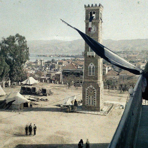 L'image montre une vue panoramique d'une place animée avec un grand bâtiment en pierre qui pourrait être une horloge ou une tour emblématique. À proximité, un soldat en uniforme se tient près de la balustrade, observant la scène. On peut voir des tentes disposées sur la place, peut-être pour un camp militaire ou une exposition. En arrière-plan, on distingue des collines et un plan d'eau, ajoutant à la profondeur du paysage. Le ciel semble dégagé, créant une atmosphère paisible.