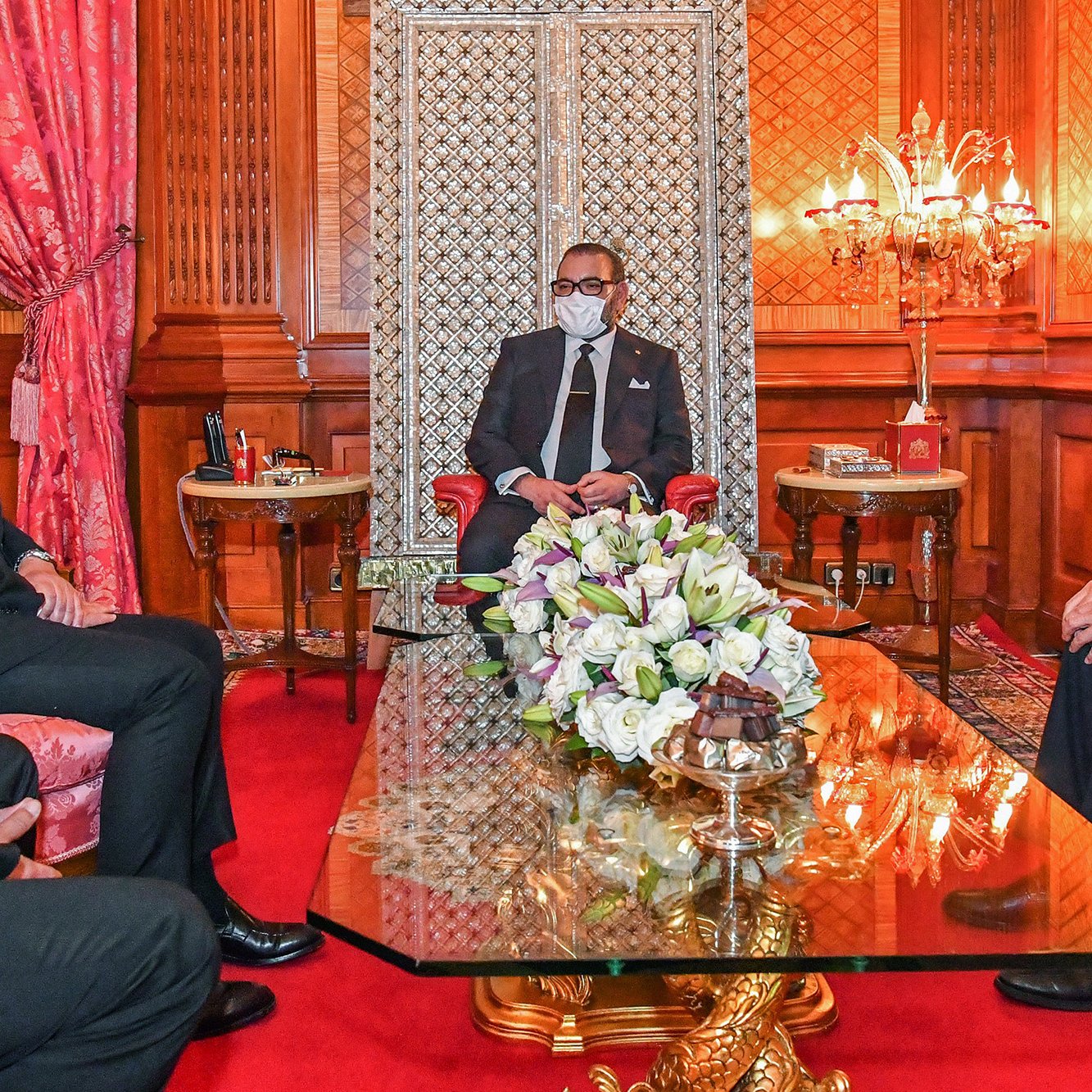 L'image montre un intérieur élégant avec un décor riche. Au centre, une table en verre repose sur un tapis rouge. On voit un homme assis, probablement un leader ou une personnalité importante, entouré de trois autres personnes, toutes en tenue formelle. Ils portent des masques, suggérant que la rencontre se déroule dans un contexte de précautions sanitaires. Le fond présente des murs ornés et un espace bien éclairé, avec des éléments décoratifs comme des fauteuils et des fleurs sur la table.