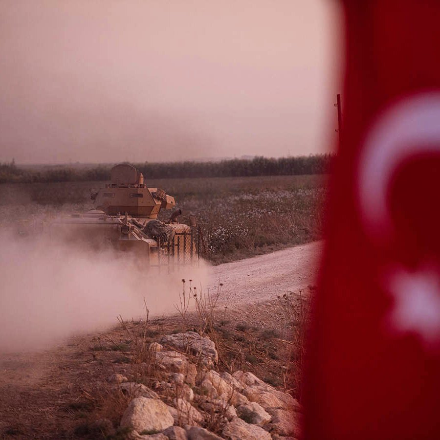 L'image montre un tank, probablement militaire, se déplaçant sur une route poussiéreuse. Au premier plan, on aperçoit un drapeau turc, qui ajoute un contexte national à la scène. L'arrière-plan présente un paysage rural, avec des champs, et l'on peut voir de la poussière se lever autour du tank, suggérant une atmosphère de mouvement ou d'activité militaire. L'éclairage est doux, créant une ambiance de crépuscule.