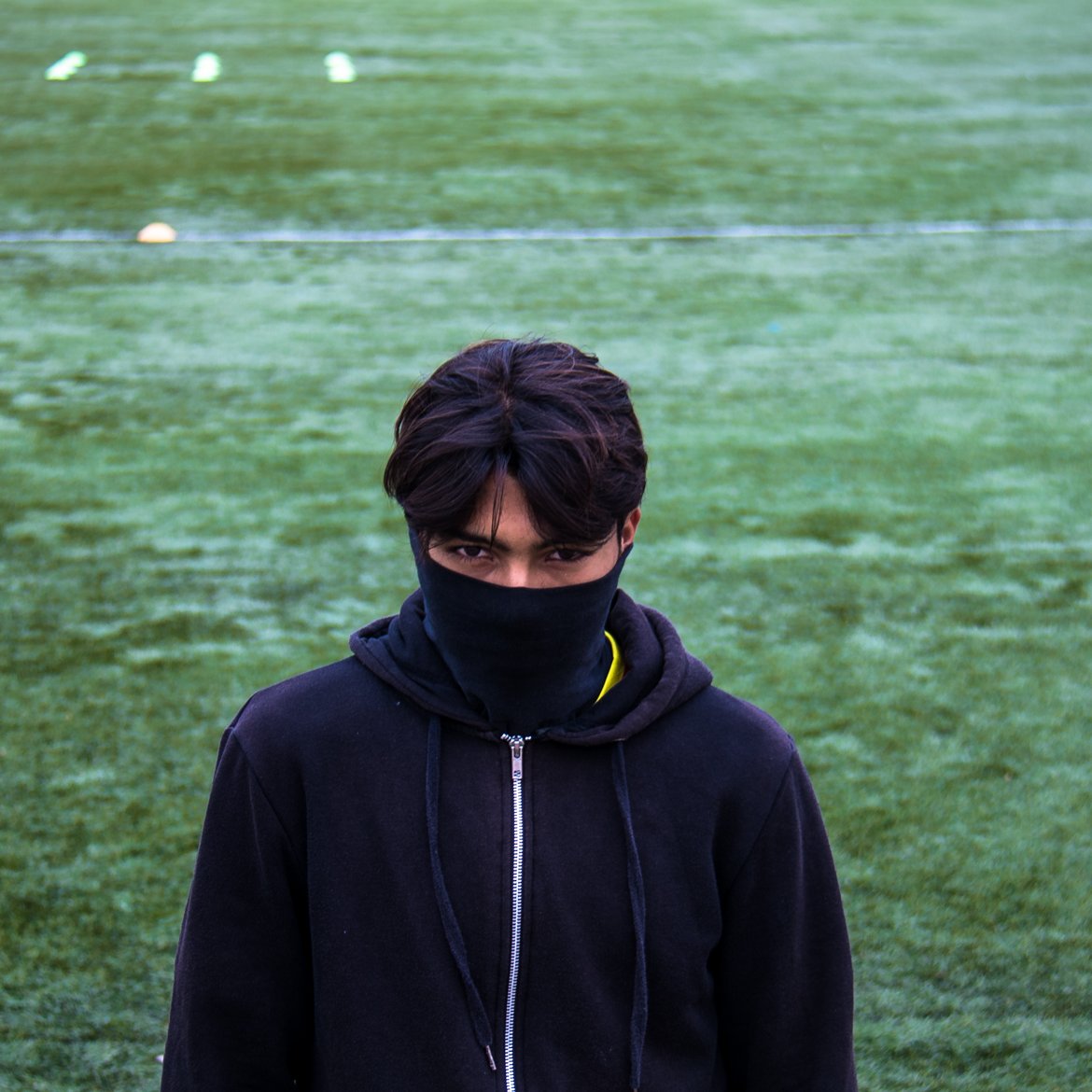 The image shows a young person standing on a sports field covered with artificial turf. They are facing the camera with a serious expression, wearing a black hoodie and a mask that covers the lower half of their face. The background appears to be a green, well-maintained playing surface with some markers on the ground suggesting it may be used for sports activities. The overall tone of the image feels intense and somewhat mysterious.