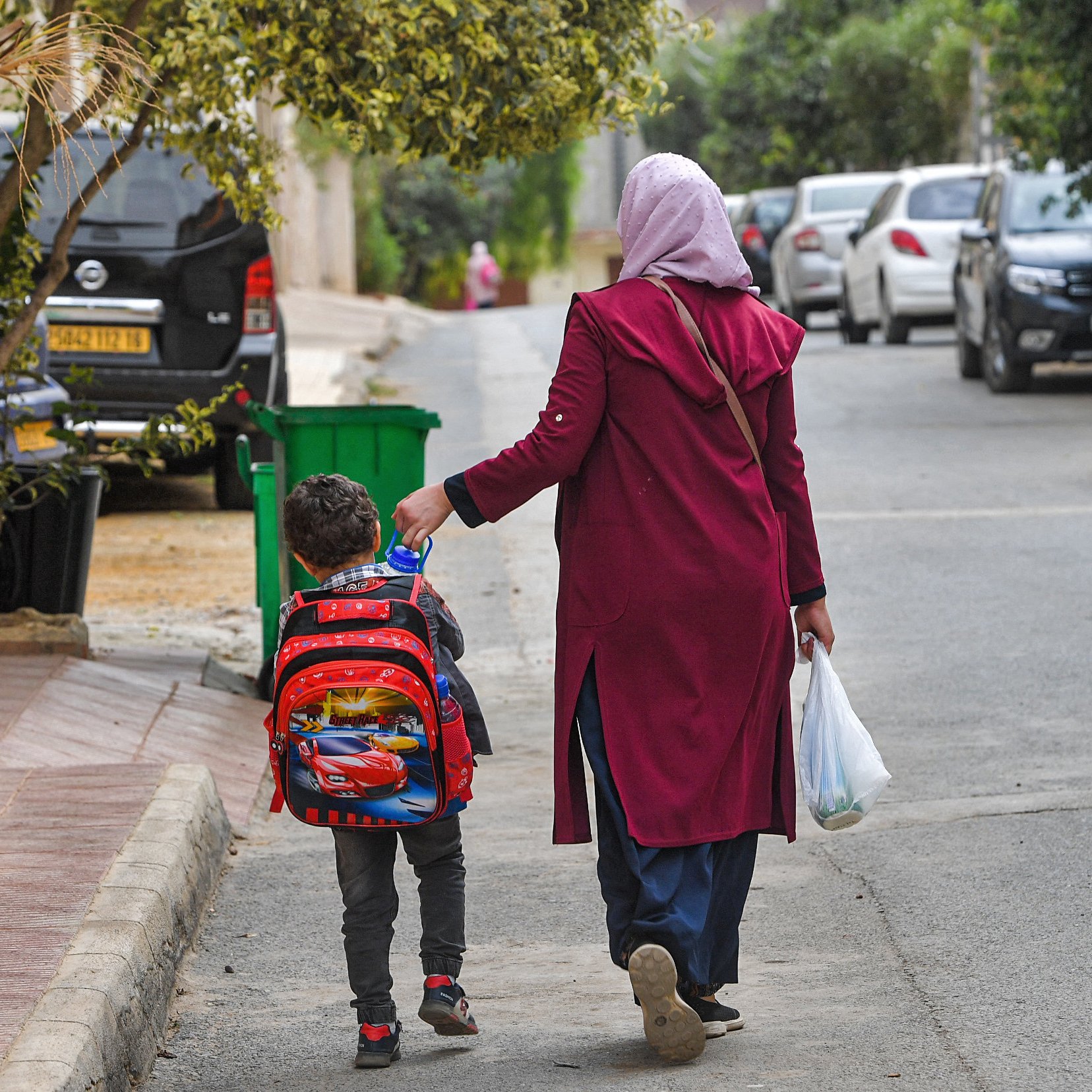 تظهر في الصورة امرأة ترتدي حجابًا وتلبس معطفًا طويلًا، تمشي مع طفل صغير يحمل حقيبة مدرسية على ظهره. يبدو الطفل سعيدًا وهو يمسك بيد والدته. الشارع محاط بالأشجار والمنازل، وهناك سيارات متوقفة على الجانبين. الأجواء تبدو هادئة وتعكس لحظة يومية من حياة الأسرة.